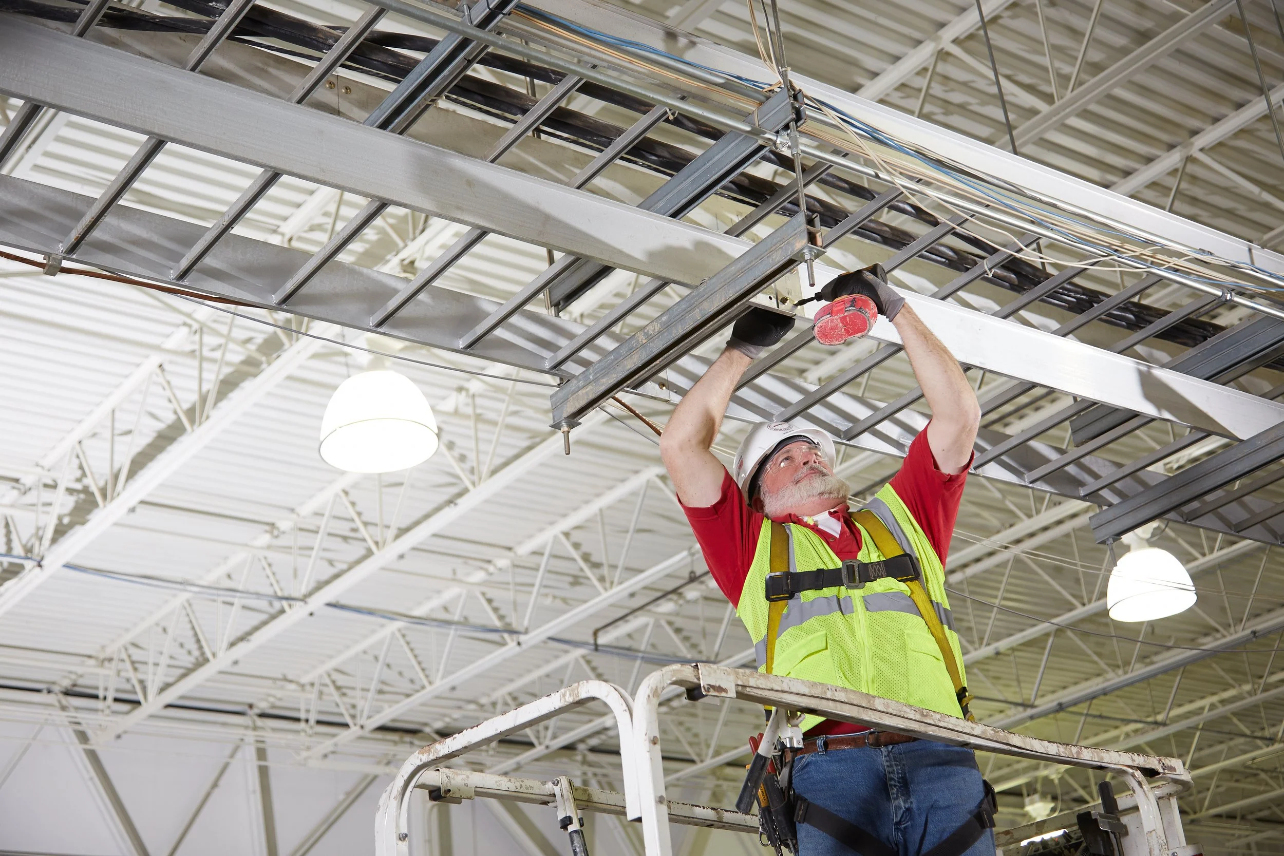 A construction worker with a helmet and safety vest repairing or installing a hanging metal ladder or cable system on the ceiling of an industrial building while standing on a lift.