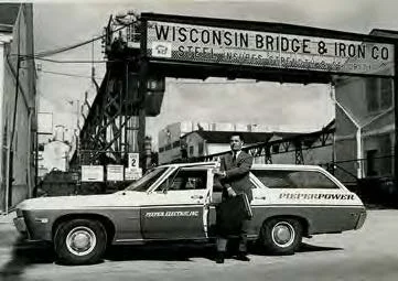 A man in a suit and hat standing next to a marked police car on a city street, with a bridge and a sign reading 'Wisconsin Bridge & Iron Co.' overhead.