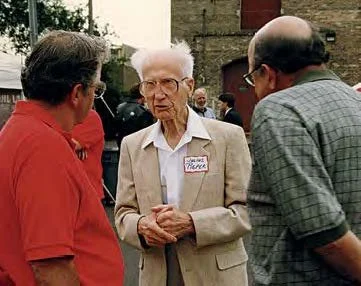 An elderly woman with white hair and glasses talking to two men outdoors, wearing a name tag, with a group of people in the background.