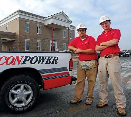 Two construction workers in red shirts and hard hats standing next to a pickup truck in a construction site, with a large building in the background.