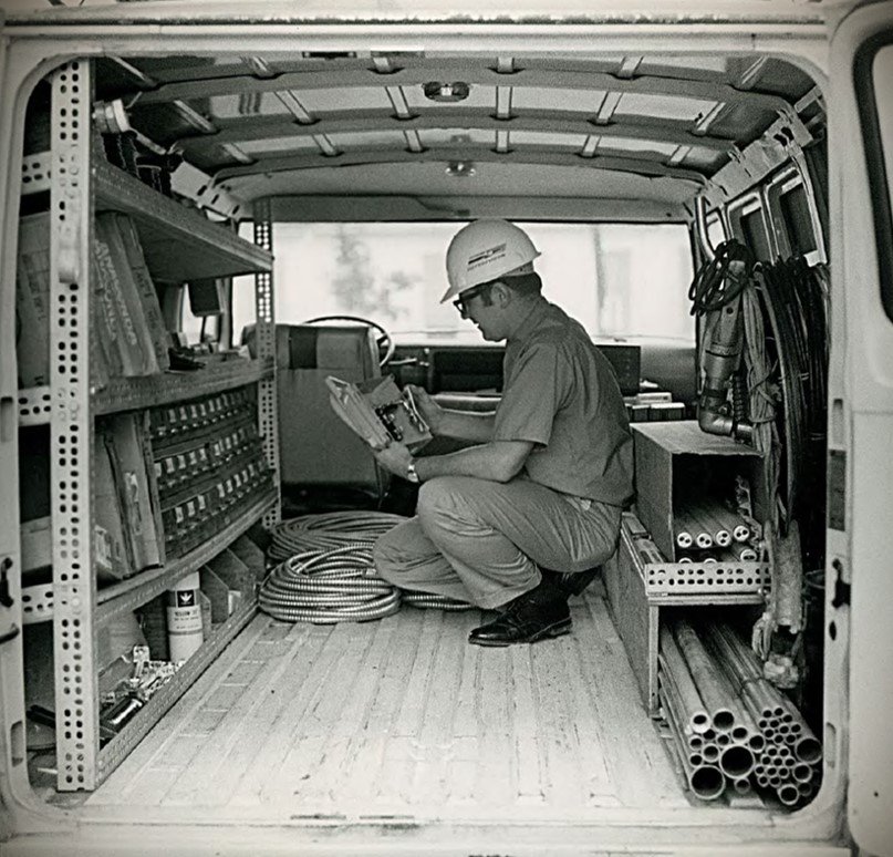 A man wearing a hard hat and work clothes is kneeling inside a utility van, surrounded by electrical equipment, pipes, and tools, inspecting or reading a device.