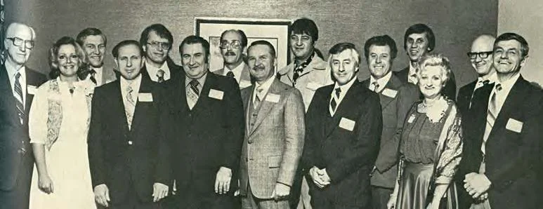 Group of 13 people, mostly men in suits, standing together indoors for a photo, with a framed picture on the wall behind them.