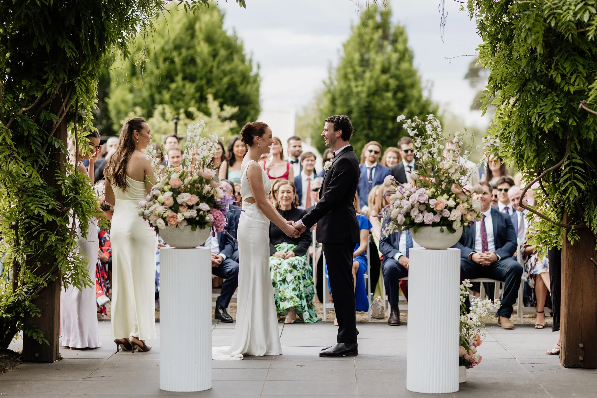 A couple getting married outdoors, facing each other and holding hands, with guests seated behind them. The scene is framed by greenery and large flower arrangements.