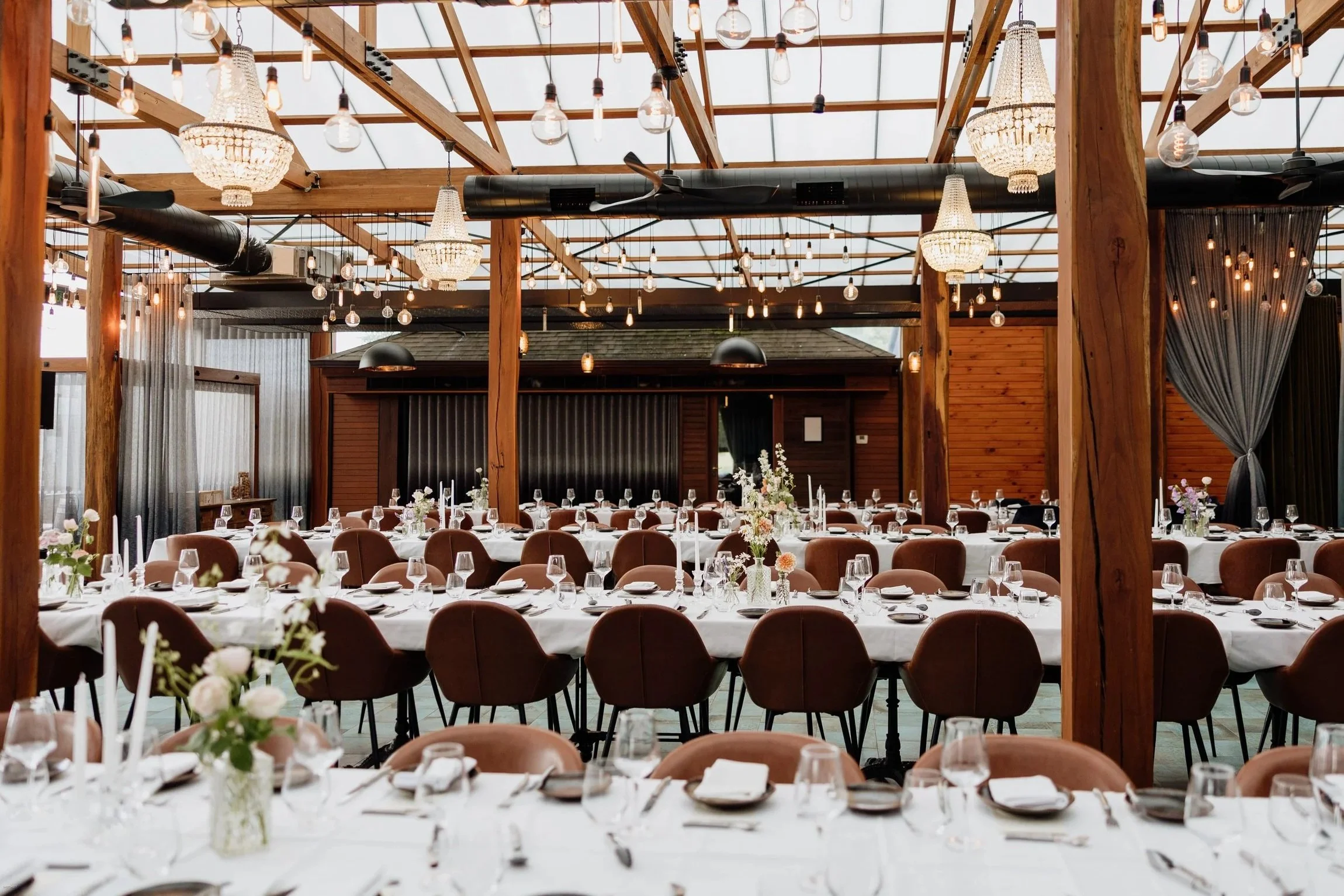 Elegant event space with long tables set for a formal dinner, decorated with flower arrangements, candles, and glassware. The ceiling has hanging light bulbs and chandeliers, with wooden beams and walls creating a rustic yet sophisticated atmosphere.