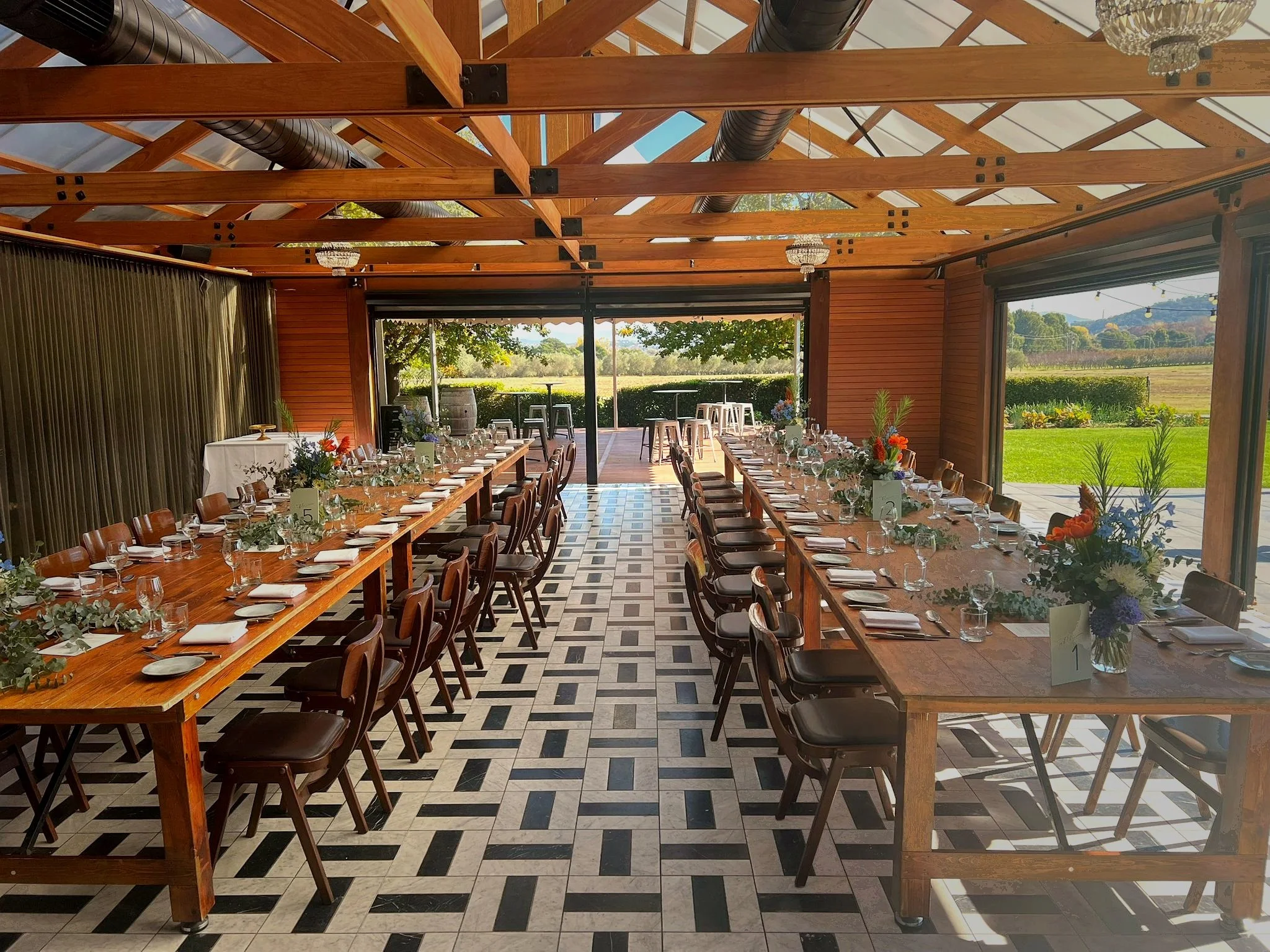 Inside a decorated banquet hall with long wooden tables set for a meal, including plates, glasses, and floral centerpieces, with large windows showing a green outdoor landscape.