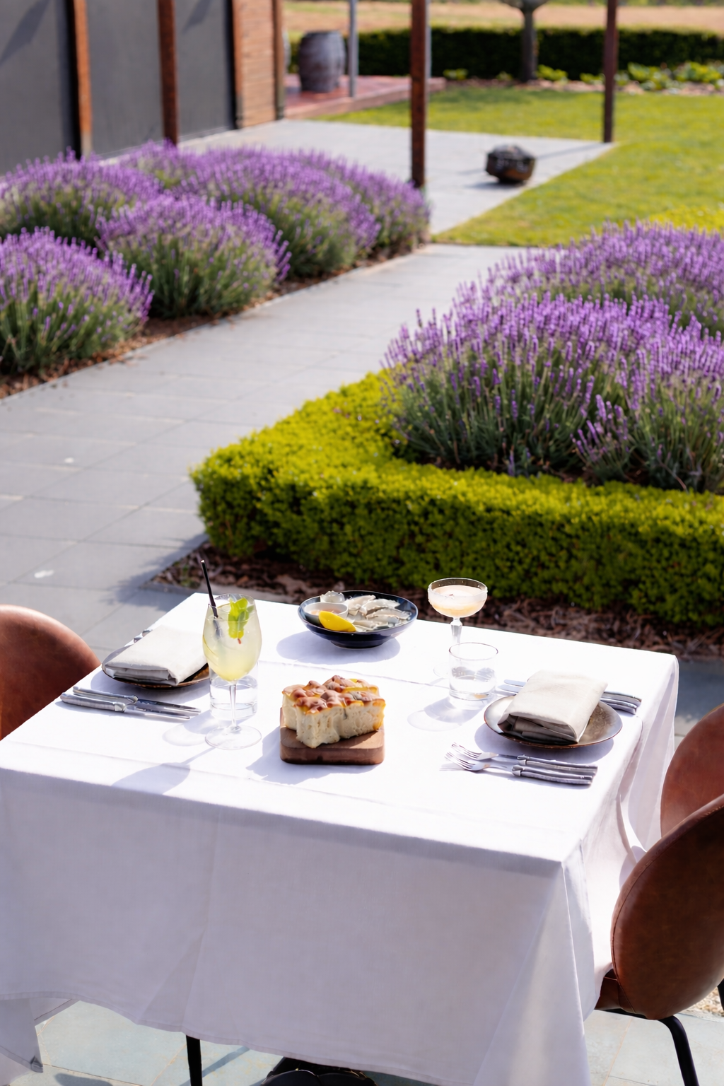 Restaurant table set for two outdoors with a white tablecloth, glasses, plates, and silverware, overlooking a garden with purple flowers and green bushes.