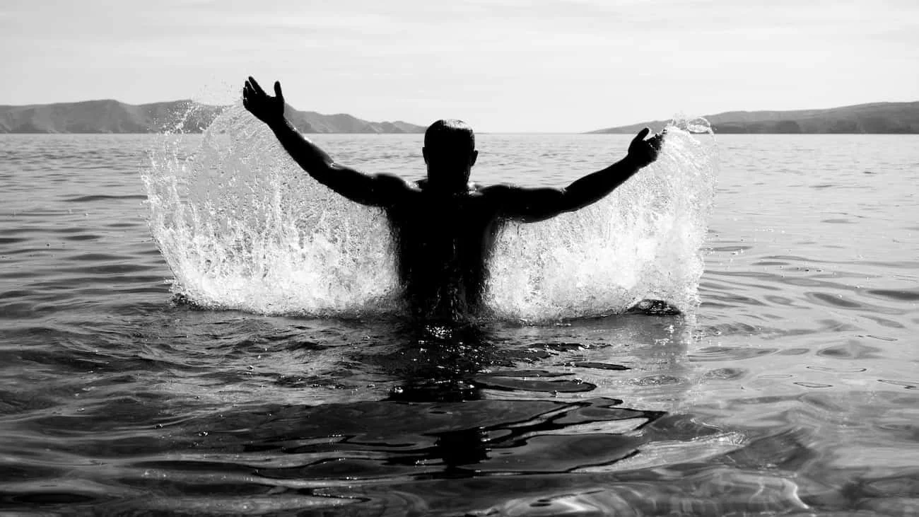 A person splashing water in a lake with arms raised, seen from behind, in black and white.