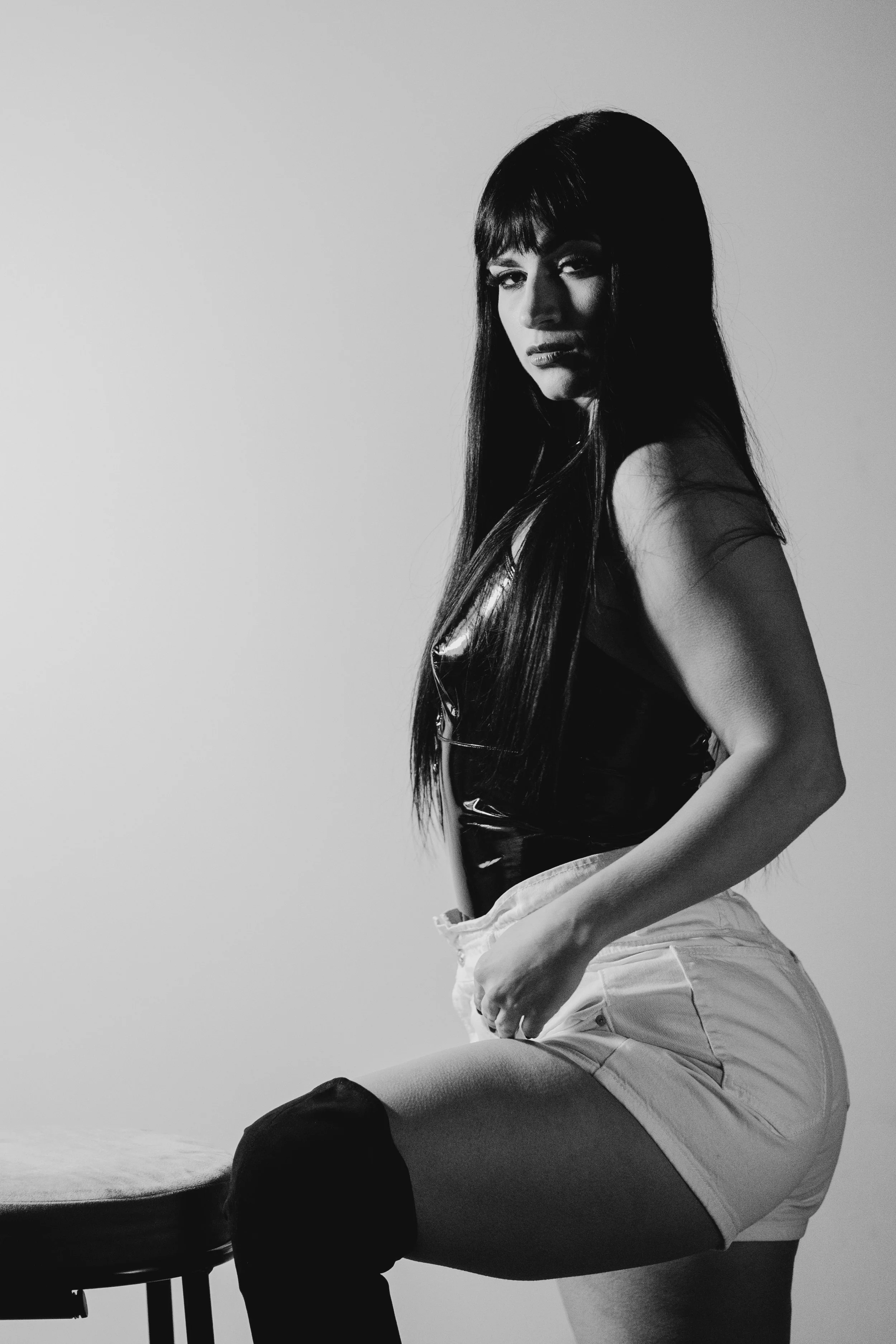 A black-and-white photograph showing a woman in a sleeveless top and shorts, sitting with one hand on her hip and the other resting on her thigh.