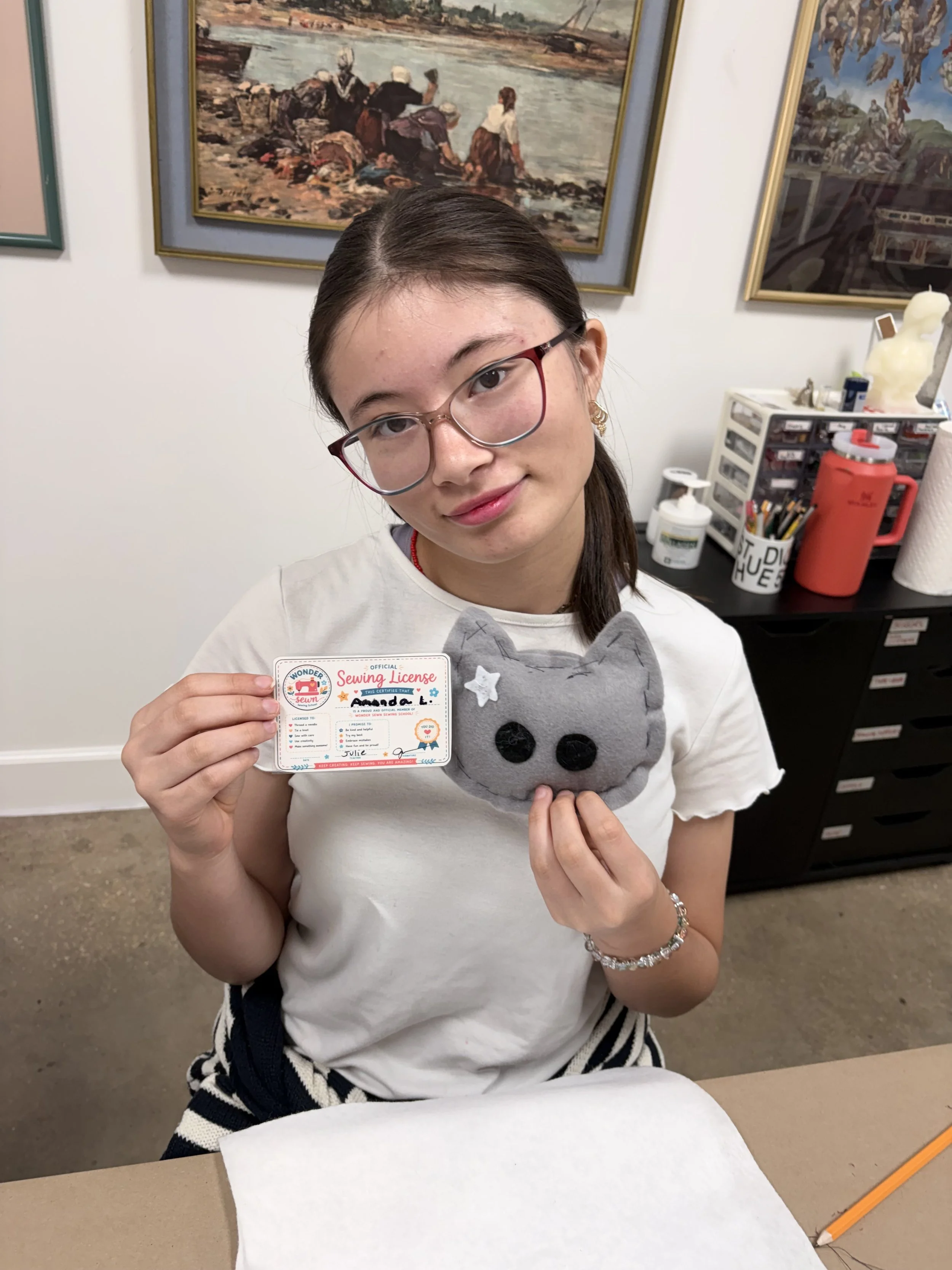 A young girl with glasses holding a sewing license and a gray plush cat toy, sitting at a table with art supplies in the background.