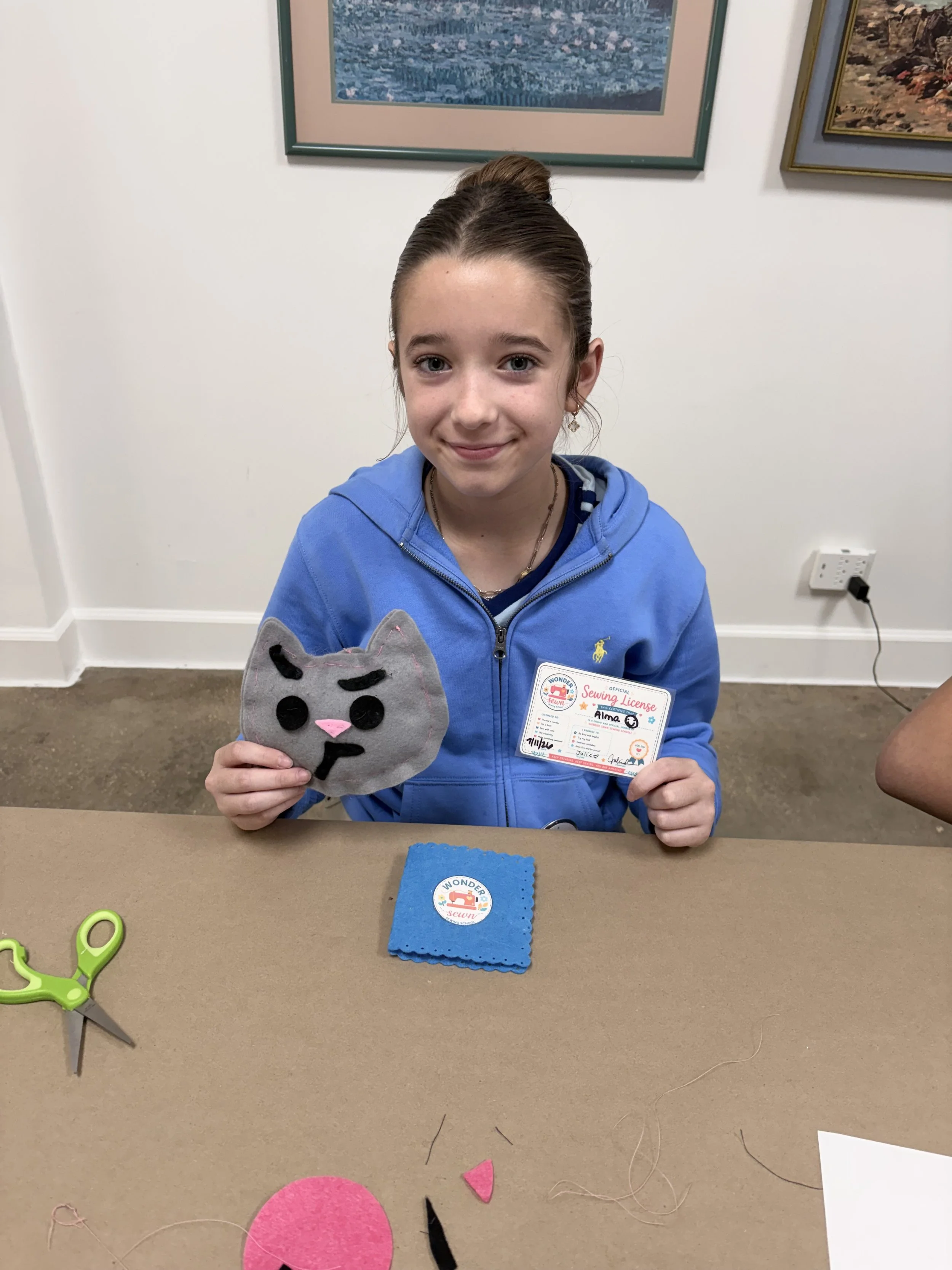 A young girl with brown hair in a bun, wearing a blue hoodie, sitting at a table holding a gray plush cat face with black eyes, nose, and mouth. She has a sewing license badge with her name Alma, and is smiling at the camera. On the table in front of her are colorful felt pieces, a blue cloth, and scissors. The background has framed pictures on a white wall and an outlet with a plug.