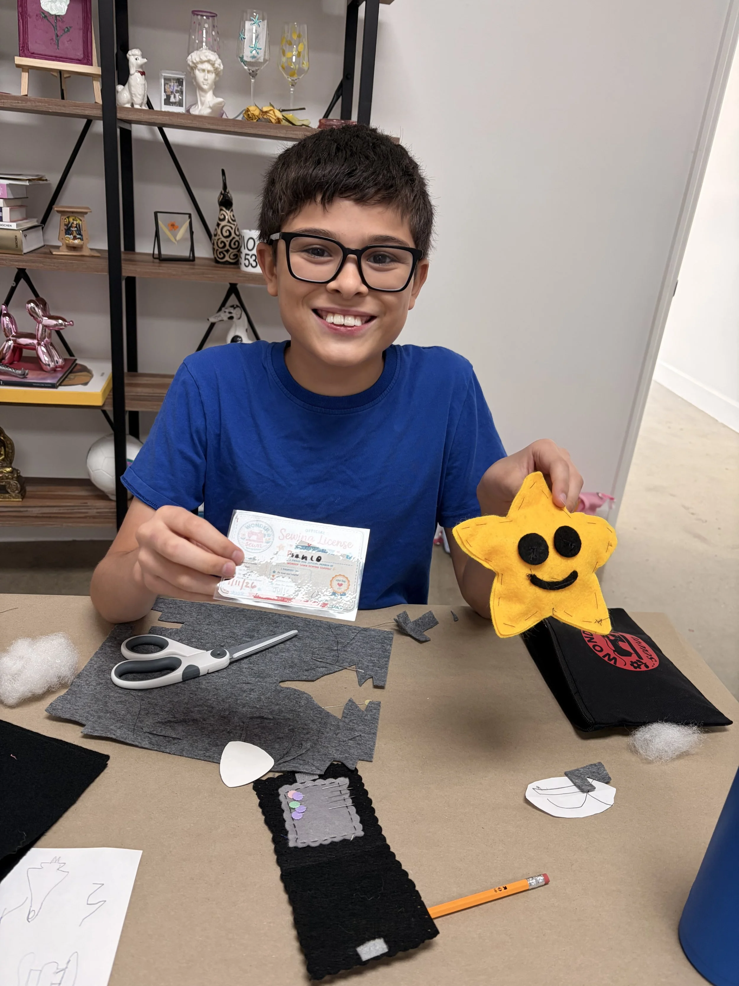 A boy with dark hair and glasses smiling while holding a yellow felt star craft with a face at a crafting table. On the table are scissors, felt pieces, and a white sheet of paper with a drafted outline. In the background, there are shelves with decorative items, books, and glassware.