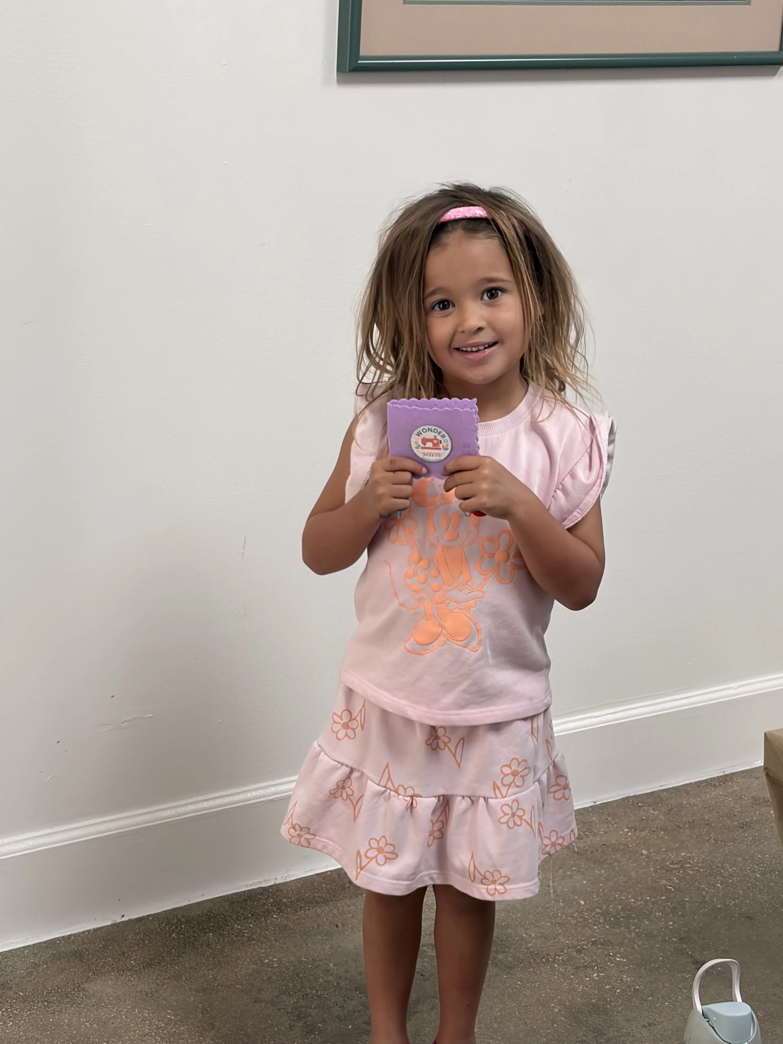 A young girl with brown hair wearing a pink dress with orange flower designs, holding a purple card, standing indoors against a white wall and brown carpeted floor.