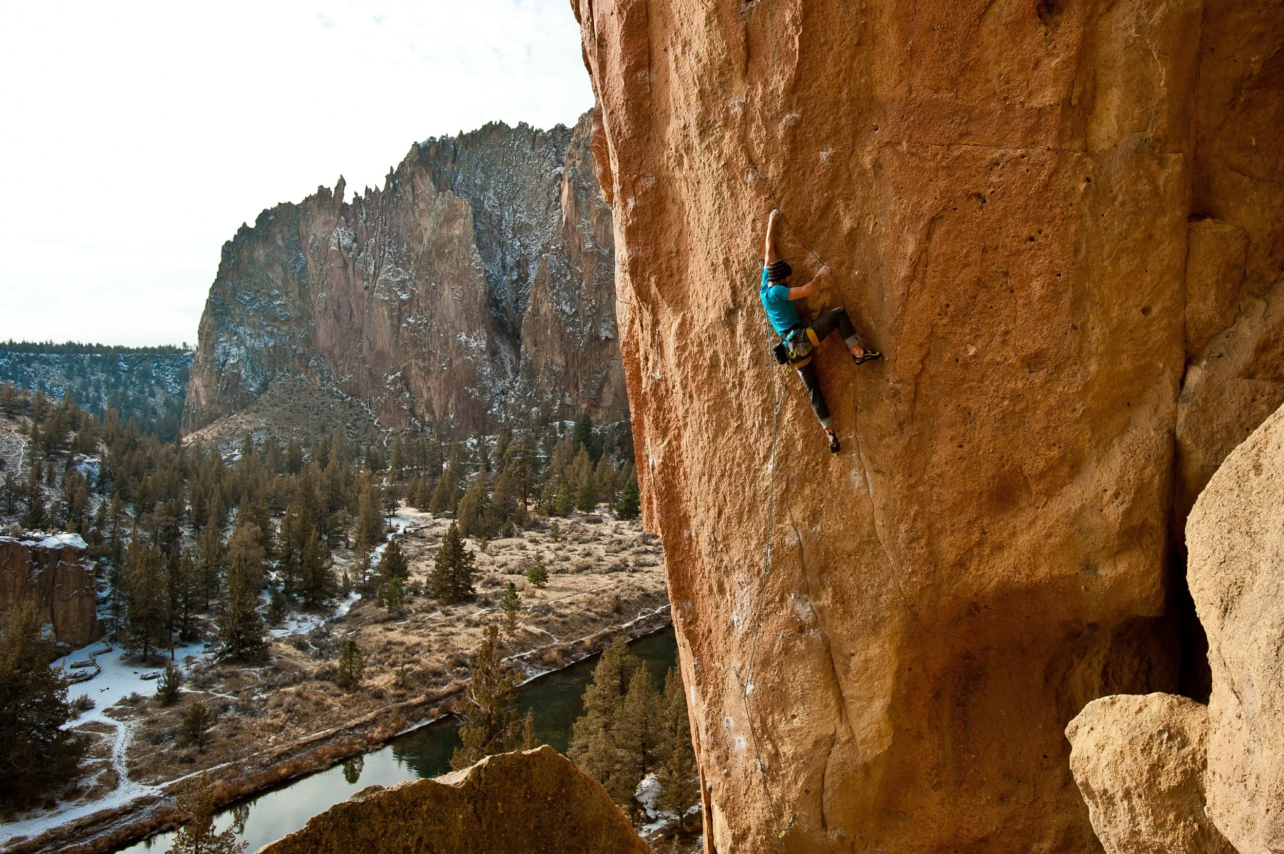 A person rock climbing on a vertical red sandstone cliff in a mountainous landscape with snow and trees, a river running below.