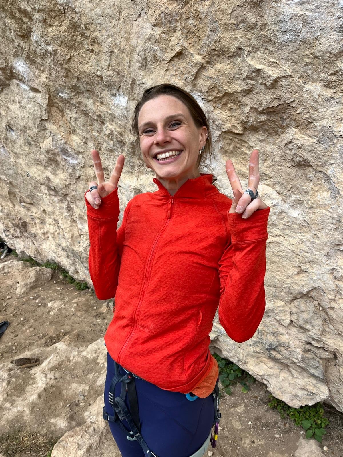 A woman in red outdoor jacket smiling and making peace signs with both hands, standing against a rocky background.