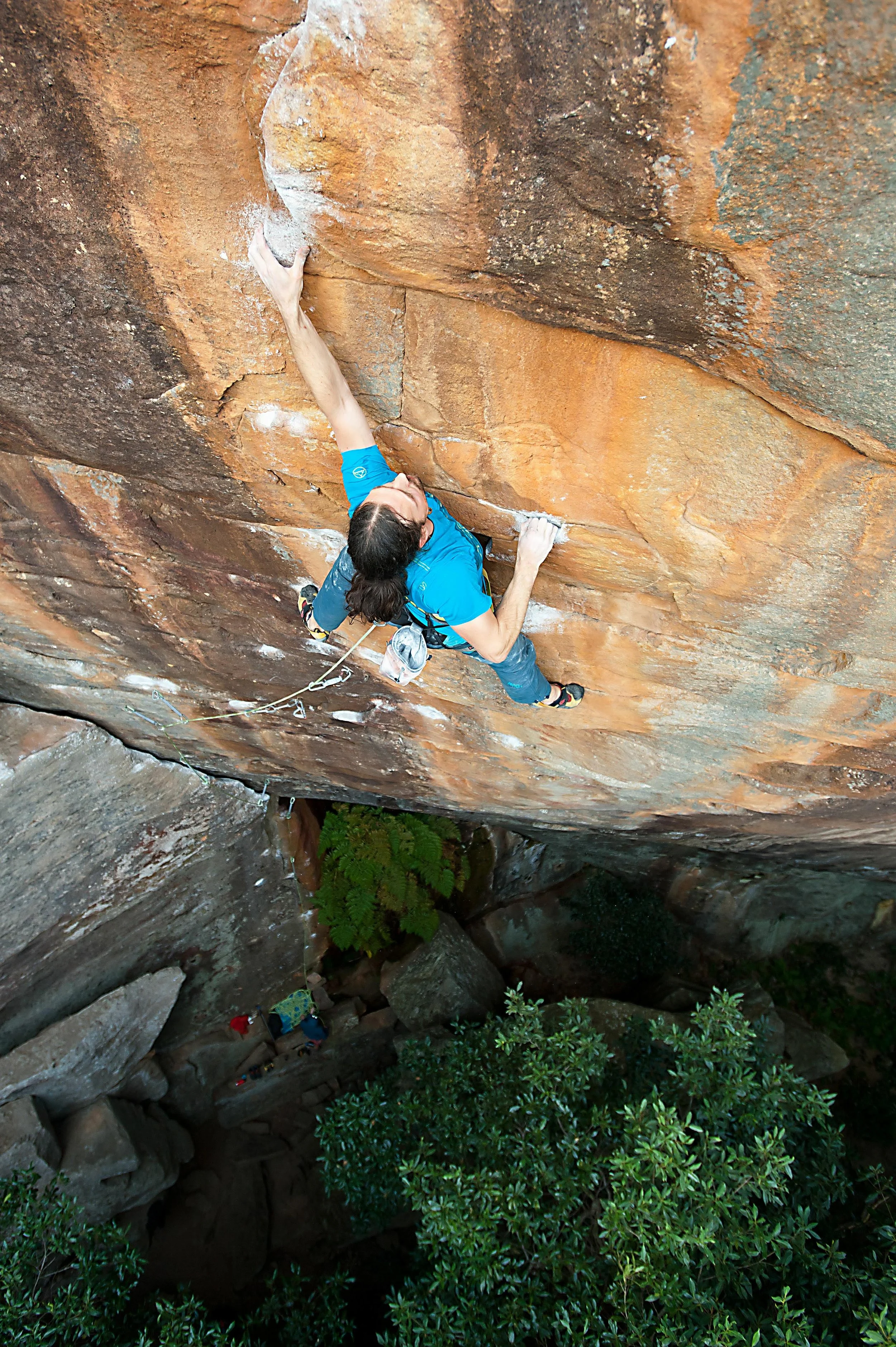 Climber in a blue shirt and jeans scaling a steep rock face outdoors, with greenery below.
