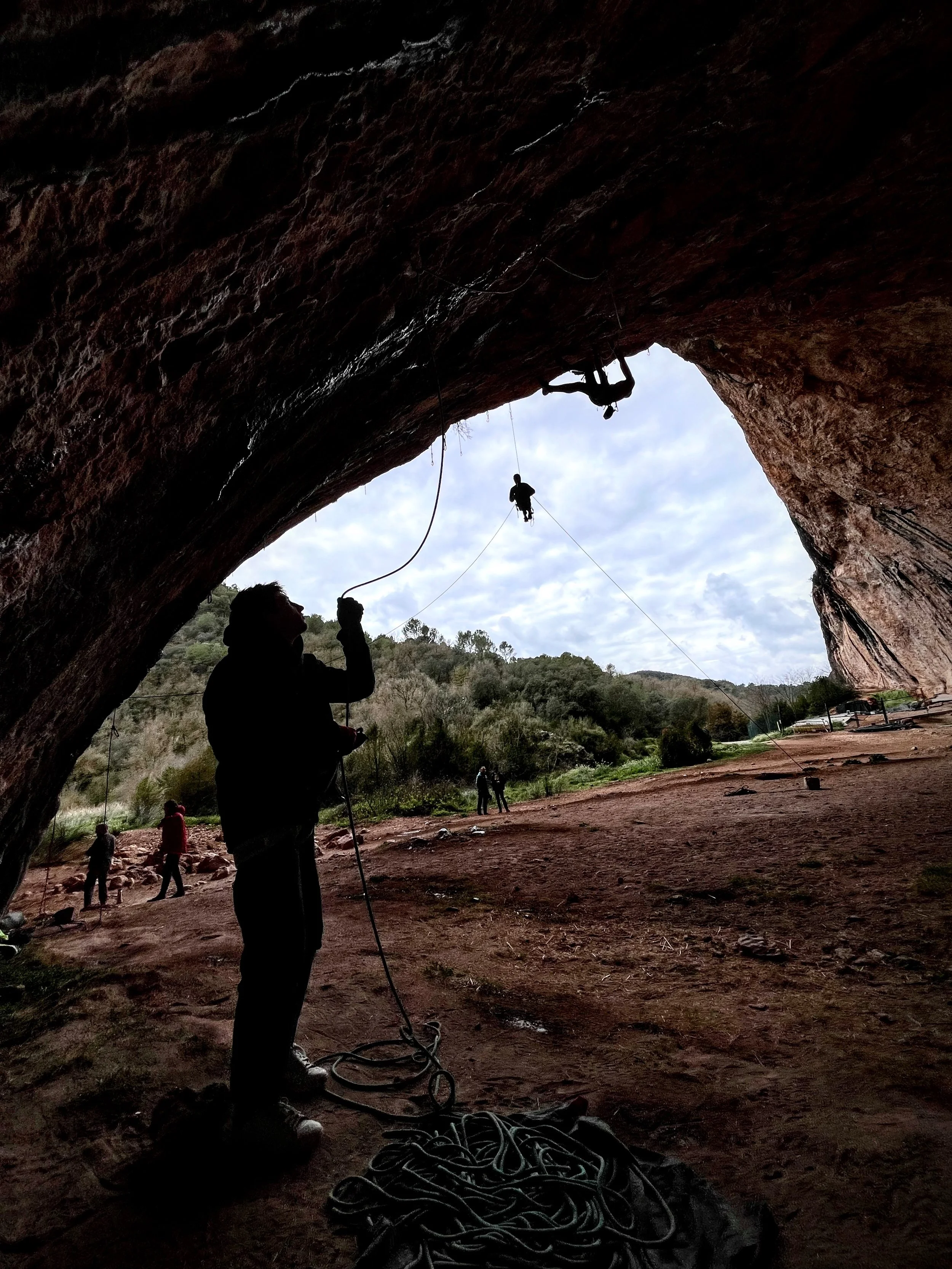 People practicing rock climbing in a cave, with one person preparing to climb and others outside the cave, under a cloudy sky.