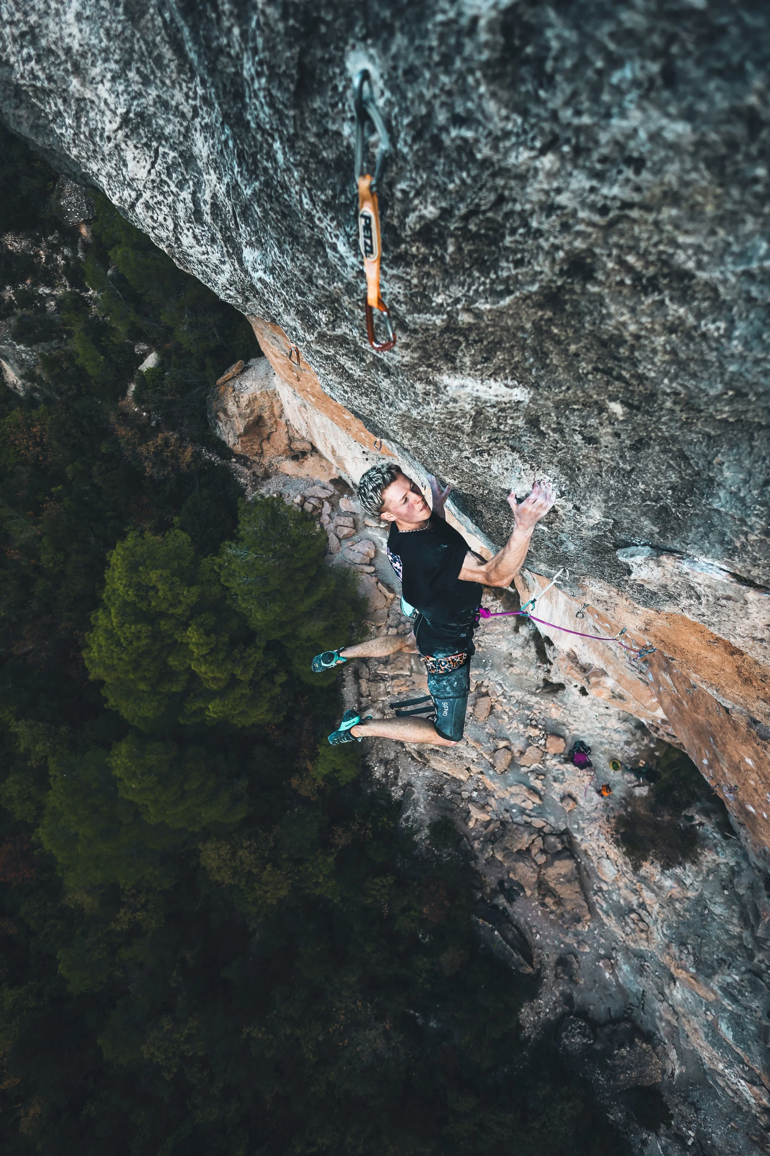 A man rock climbing on a steep cliff face, wearing climbing gear, his right hand gripping a hold, and his left hand reaching for another hold, with a safety harness attached to the rock, and other climbers below on the ground.