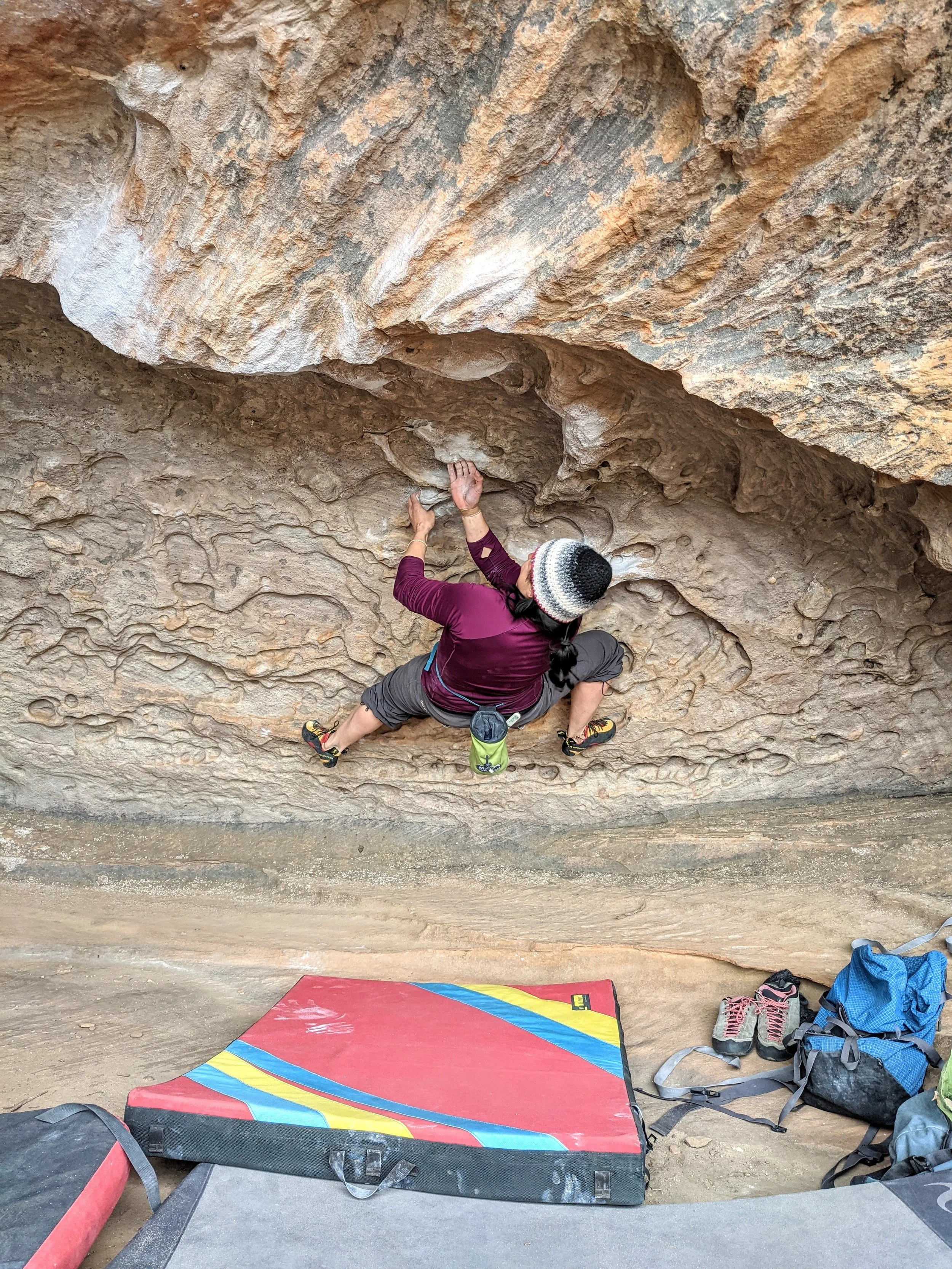A person is bouldering on an indoor rock climbing wall, wearing a striped beanie, maroon jacket, and gray pants. Climbing gear and shoes are on the ground below, along with a padded crash pad for safety.