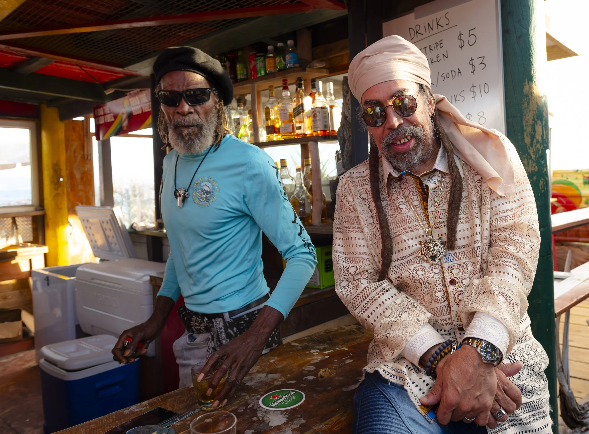 Two men with dreadlocks and sunglasses standing behind a bar, one wearing a turquoise long sleeve shirt and the other wearing a light-colored lace shirt and headscarf, with bottles of alcohol and a drink on the bar.