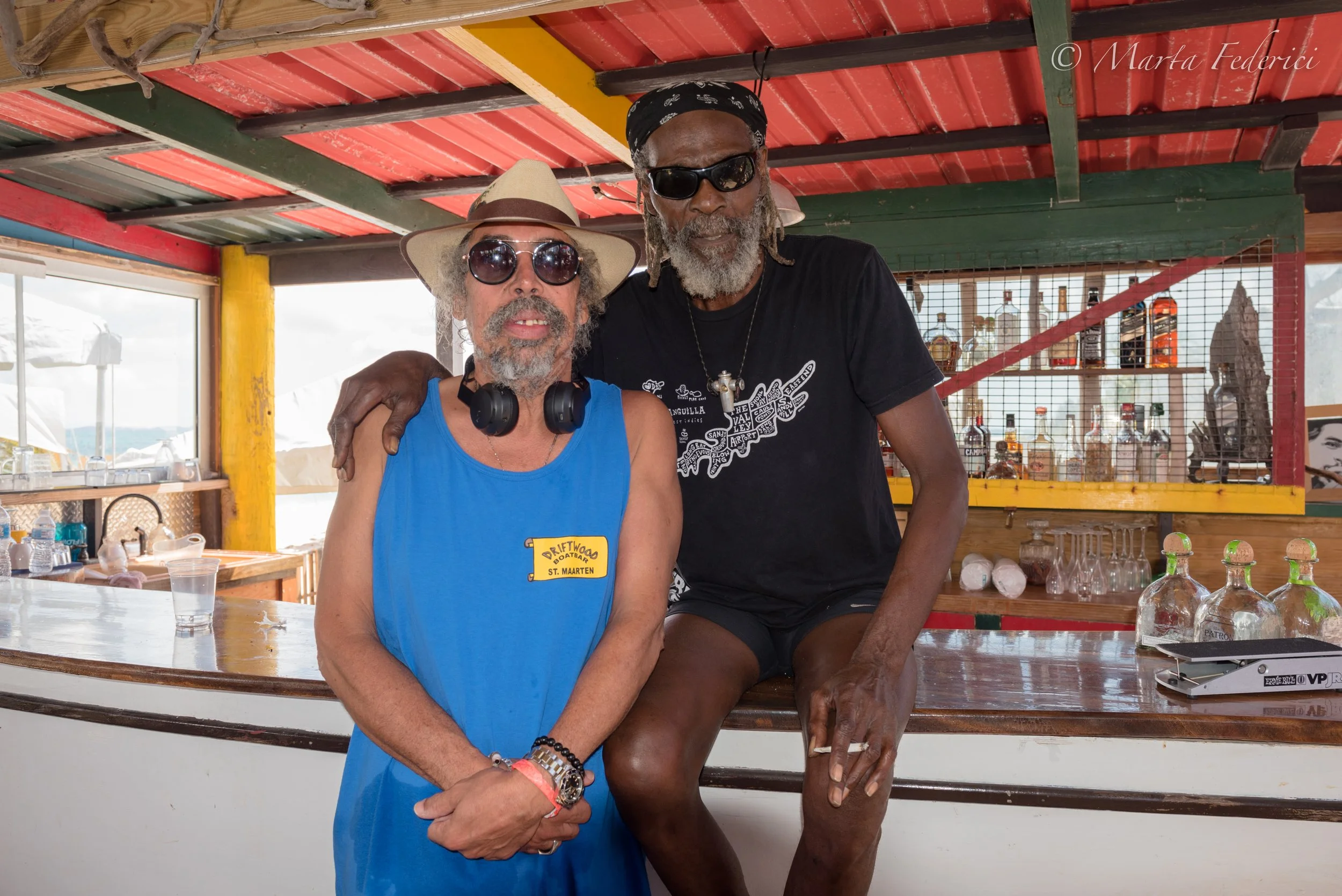 Two men with gray beards and sunglasses standing behind a bar, one with a black bandana and the other with a straw hat, posing for a photo on a boat or beachside bar, with bottles and glasses in the background.