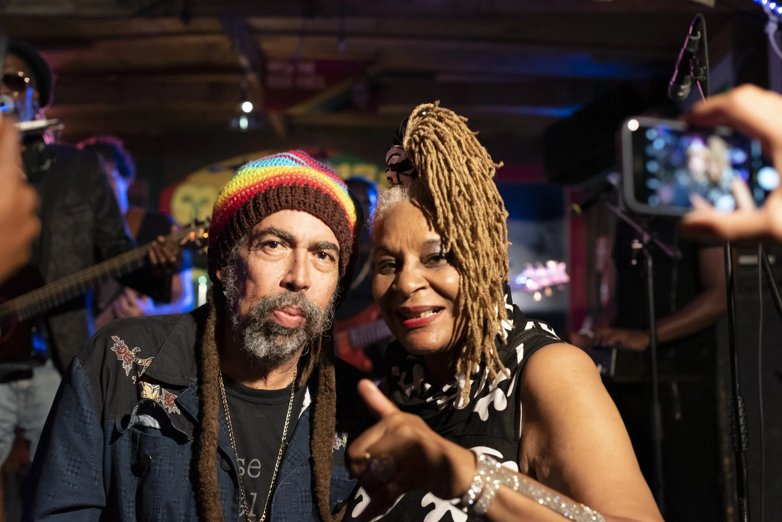 Two people posing for a photo in a lively music venue, one wearing a rainbow-colored knit hat and the other with long, blonde dreadlocks, with a band performing in the background.