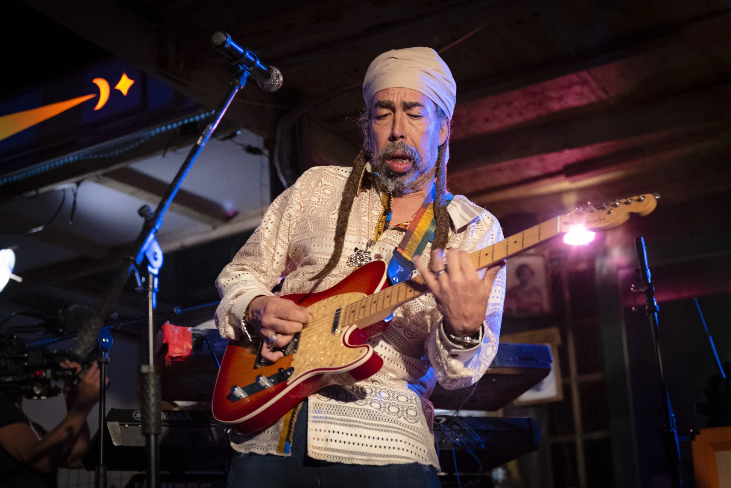 A man with dreadlocks and a white turban playing an electric guitar on stage, with a keyboard and microphones nearby.