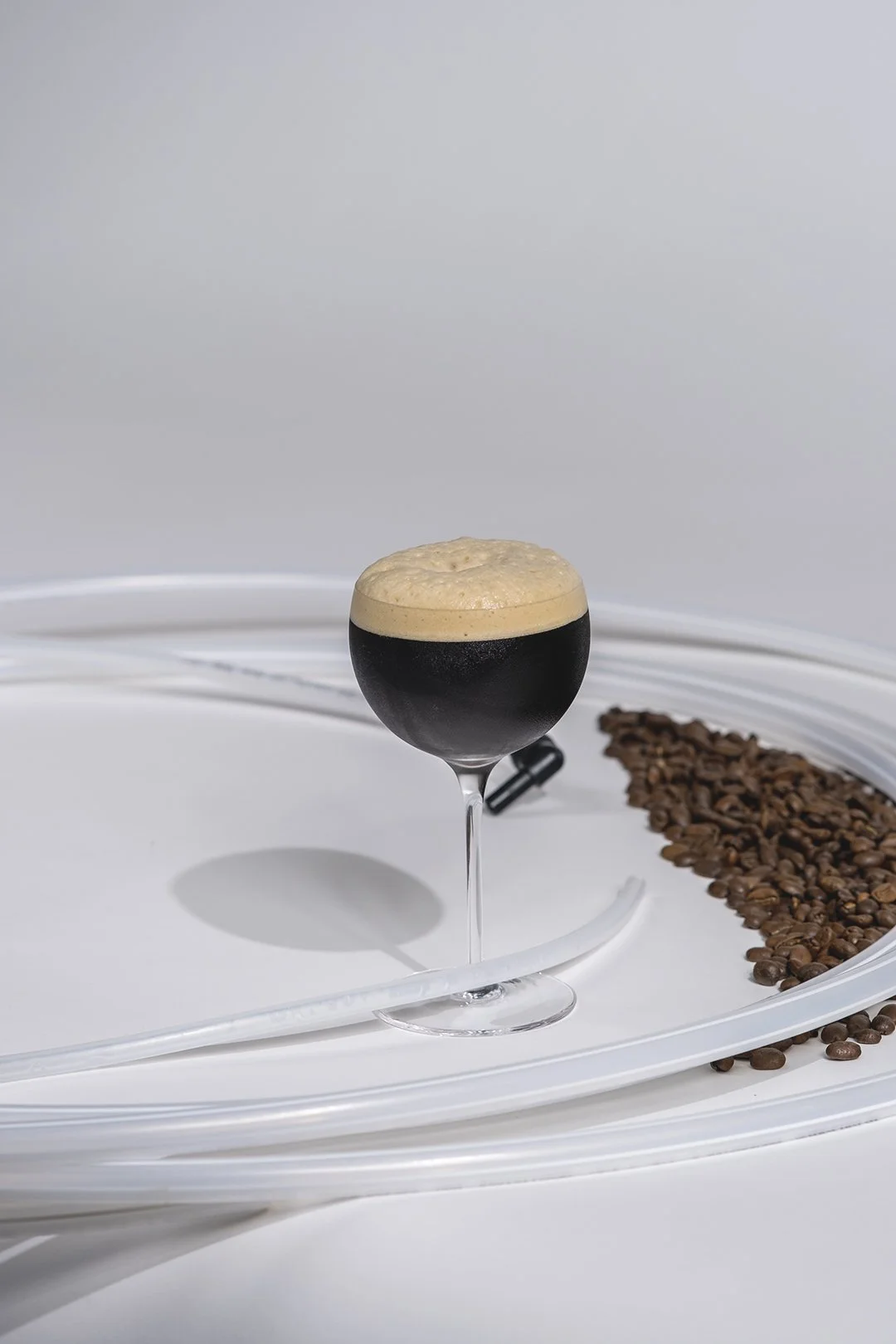 En svart öl i ett fotoglas med skum på toppen, placerad på en vit yta med en linje av kaffebönor i bakgrunden.