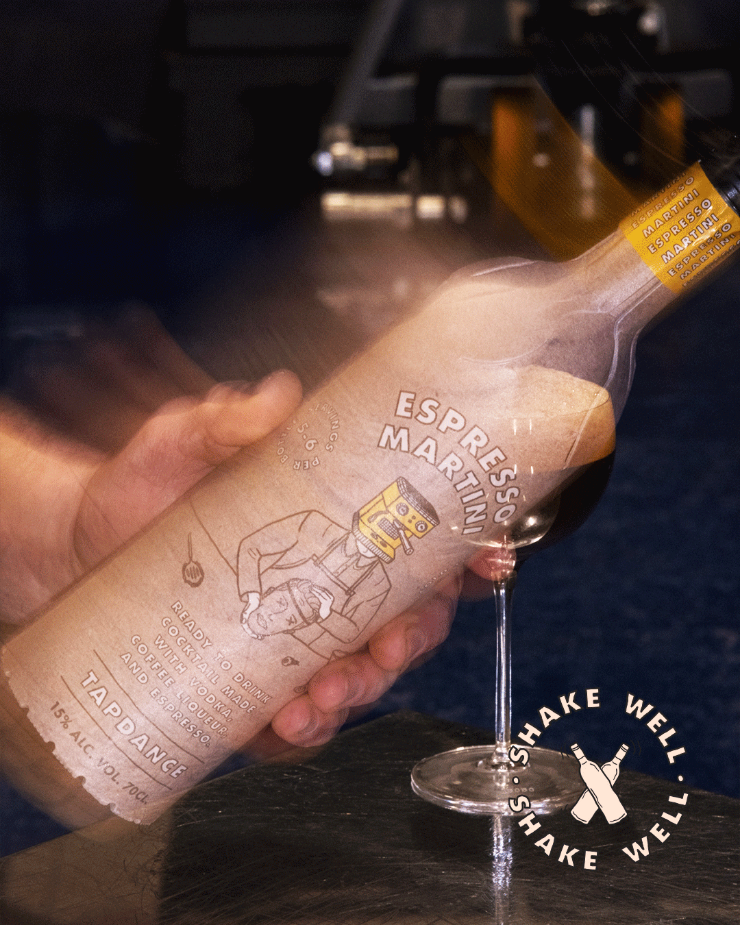 En hand som håller en flaska espressoshots som är placerad ovanpå ett cocktailglas. Flaskans etikett är i papper och har en tecknad bild av en person med en tehuvud och texten 'Espresso Martini'. Det finns även instruktioner och alkoholprocent på etiketten.