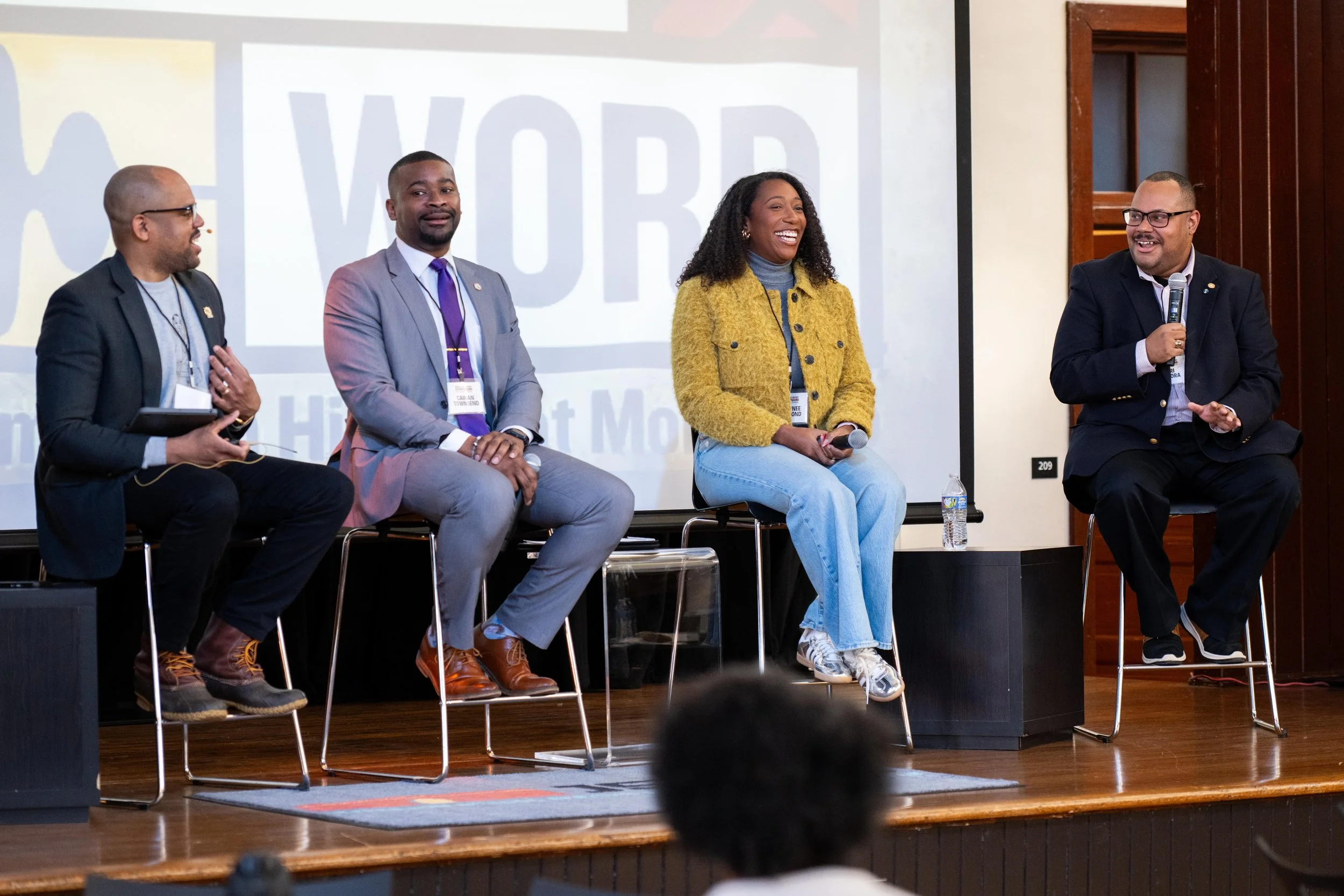 Five people sitting on stage, participating in a panel discussion. They are smiling and engaging with the audience. A woman in a yellow jacket is holding a microphone, and a man on the right is speaking into a microphone. Behind them, a large screen displays the word 'WORL'.