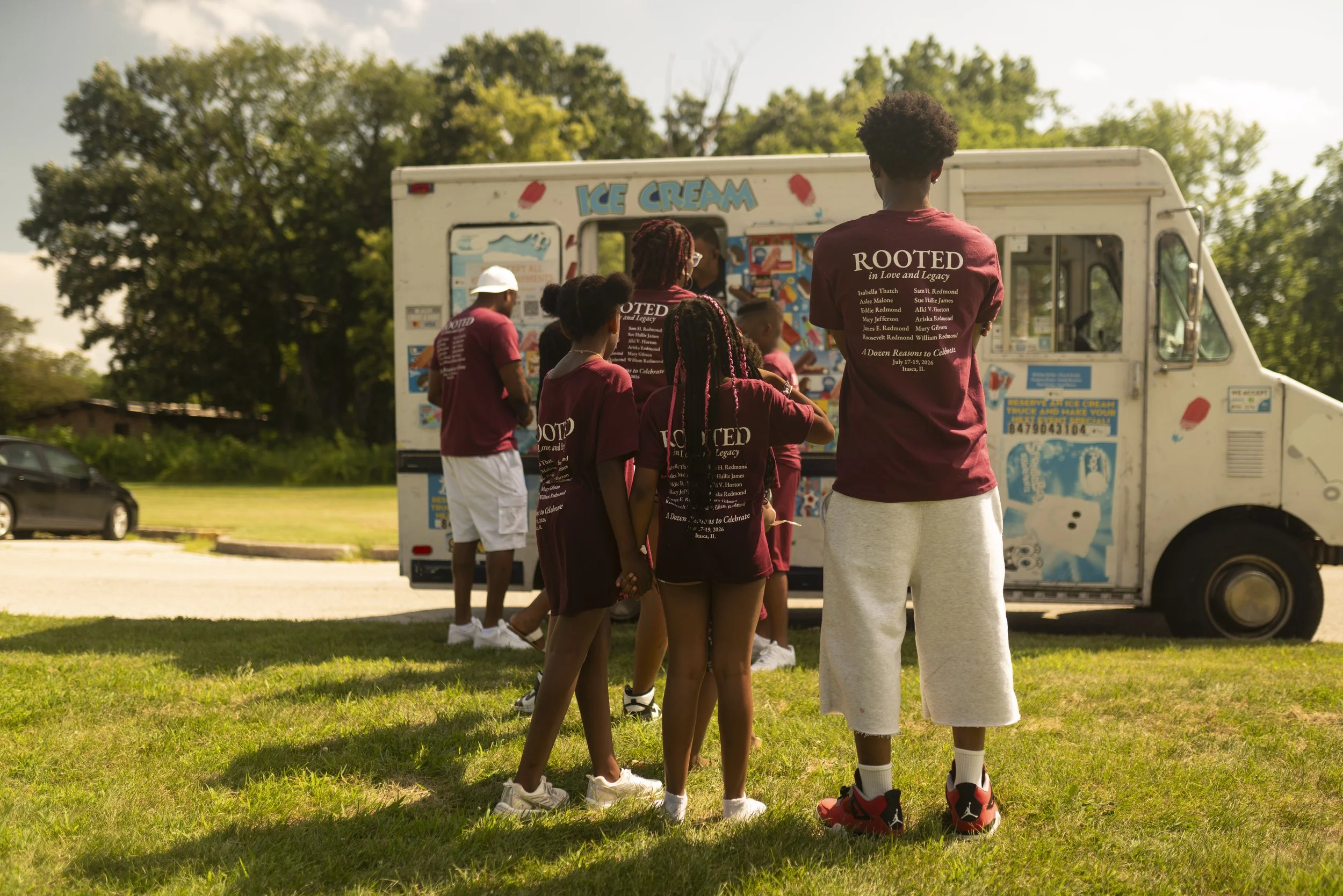A group of people, mostly children, standing in line to get ice cream at a truck. They are outdoors on a sunny day with green trees in the background. Most are wearing maroon shirts with white text.