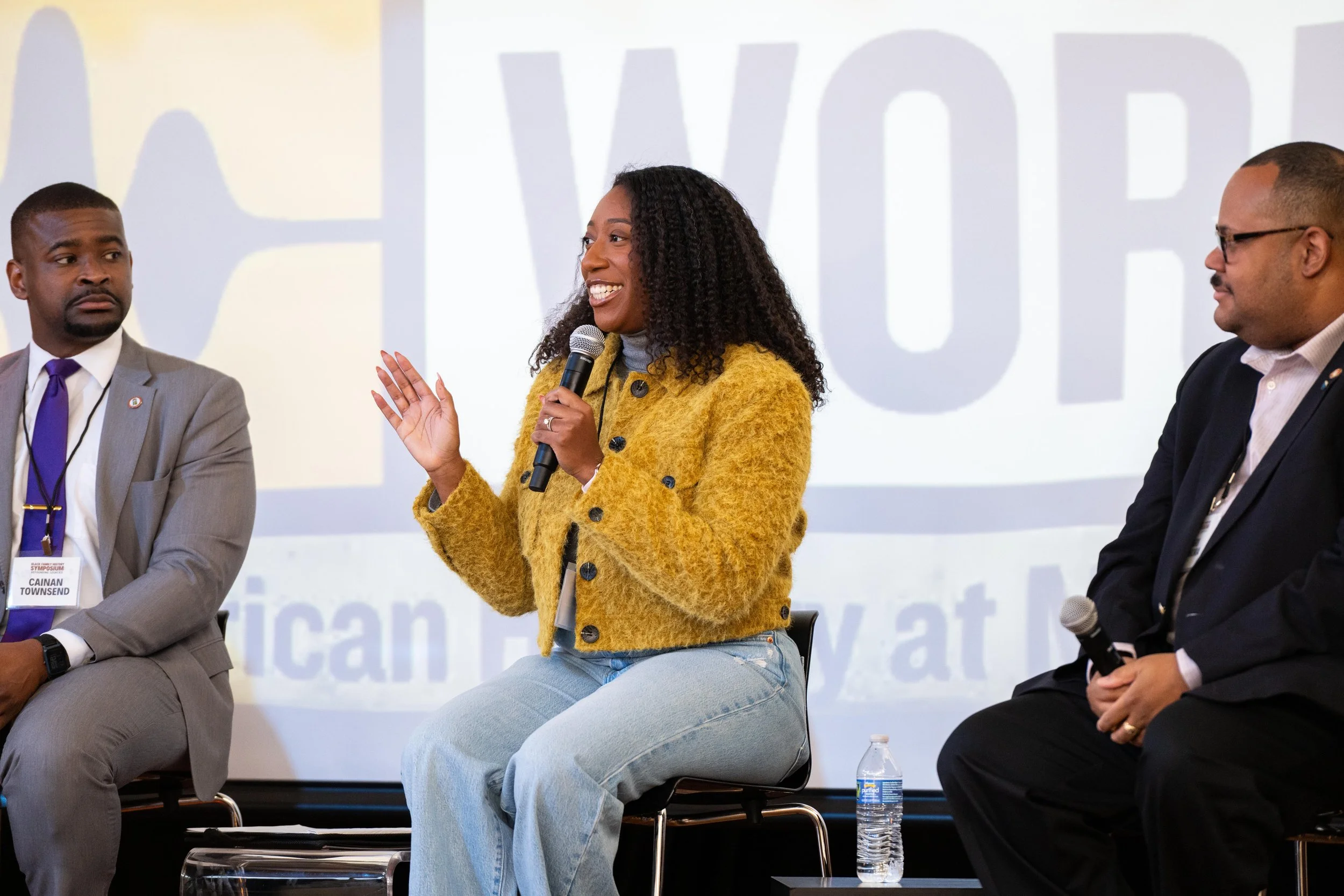 A woman in a yellow coat speaking into a microphone during a panel discussion, with two men sitting on either side and a large projection screen in the background.
