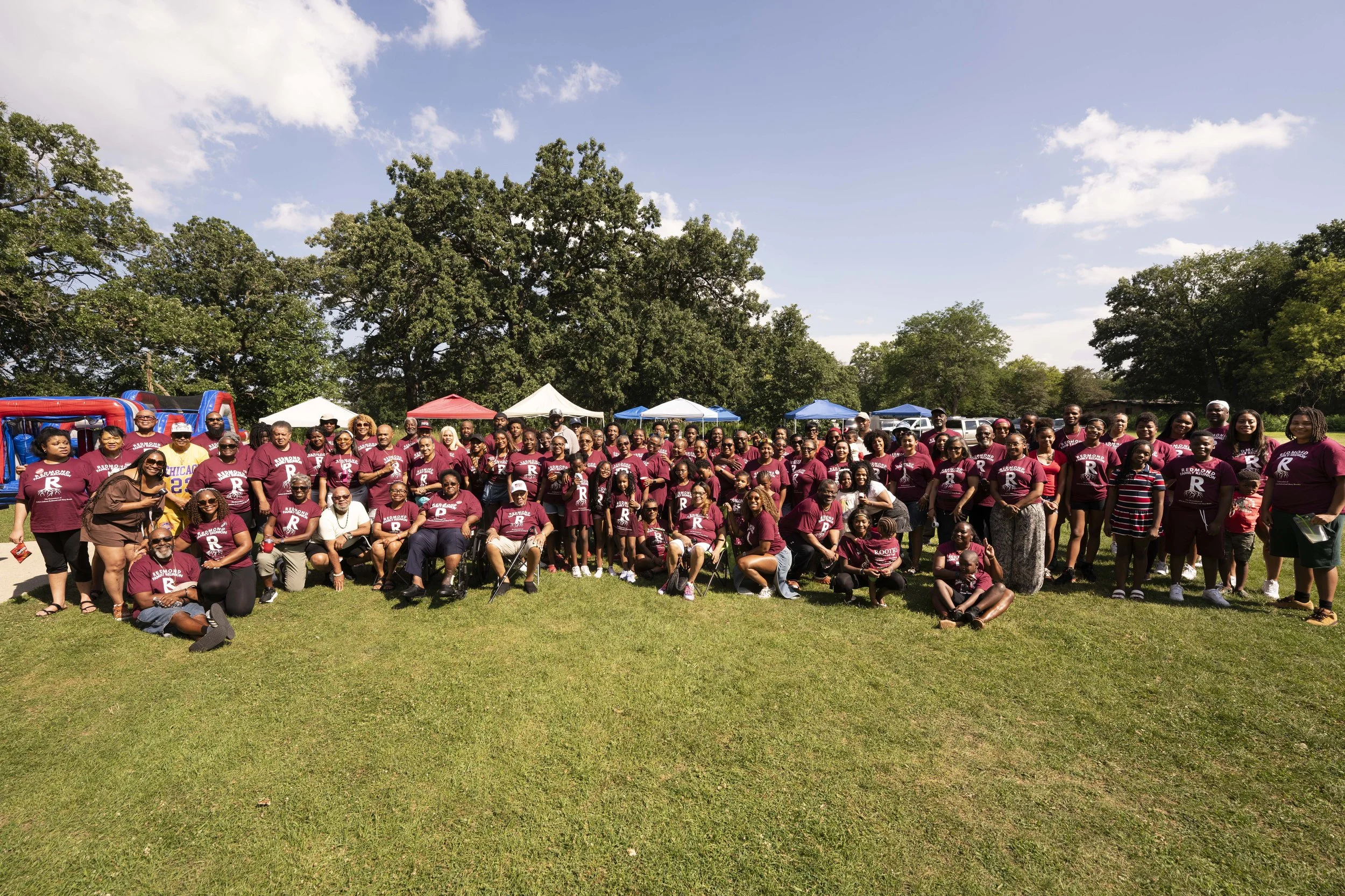 Group of people gathered outdoors for a celebration or event, many wearing matching maroon T-shirts with a large letter R and text, with tents and inflatable structure in background, under a partly cloudy sky.