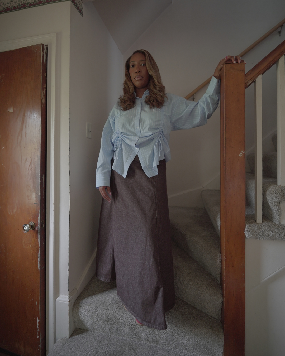 A woman standing at the bottom of a staircase holding the wooden railing, wearing a light blue blouse and a long brown skirt, in a home interior.