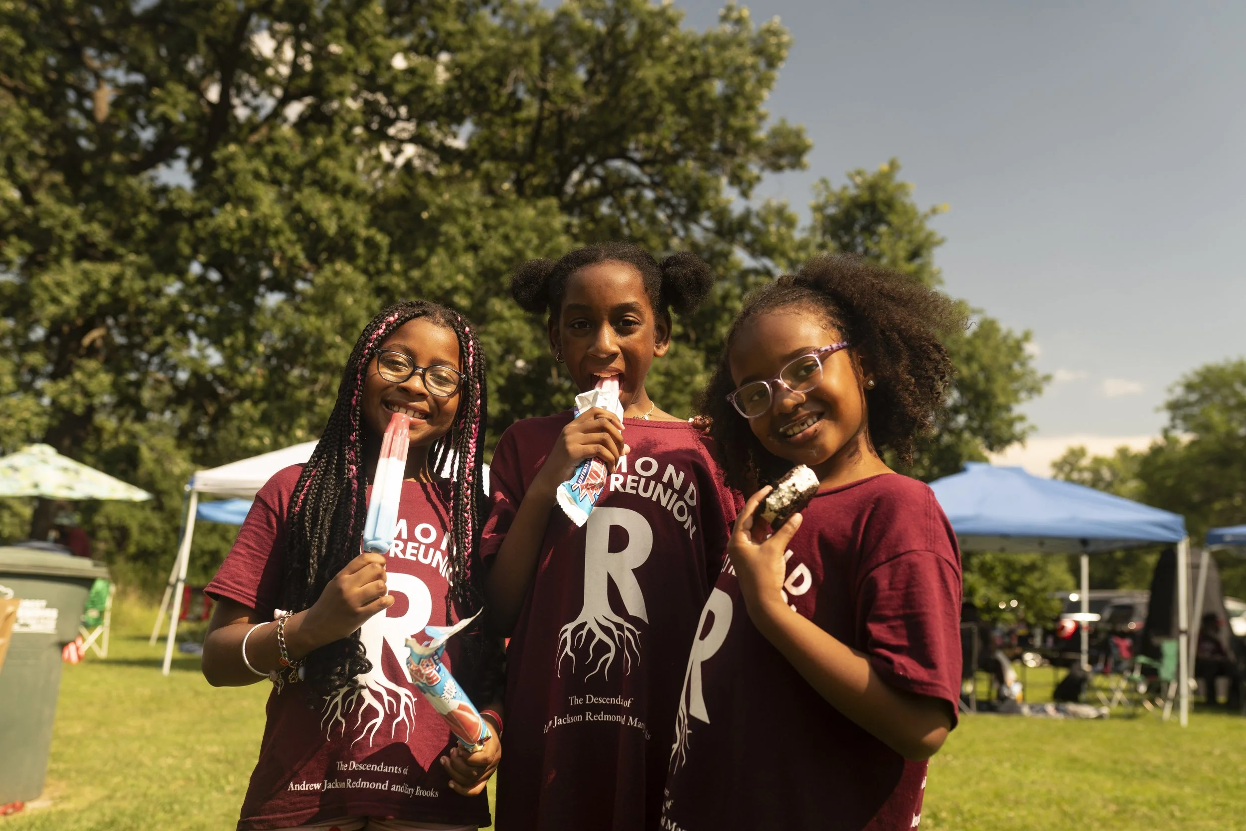 Three young women are outdoors in a park with trees and tents in the background, enjoying ice cream and smiling at the camera. They are wearing matching maroon shirts with white text and graphics.