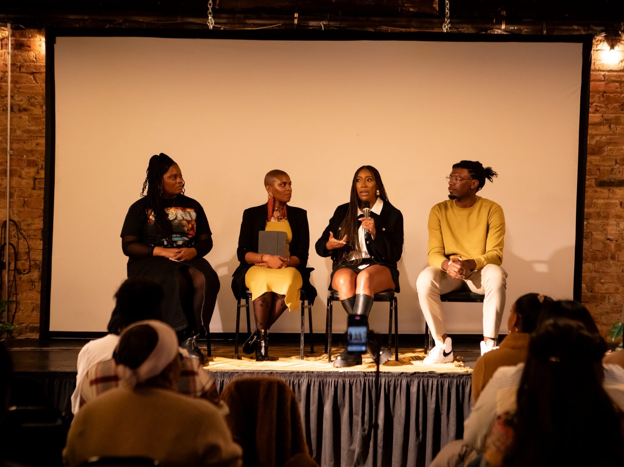 Four women sitting on chairs on a stage, participating in panel discussion, with one woman speaking into a microphone. Audience members are visible in the foreground, and a large white screen is behind the panelists. The stage has a brick wall background.