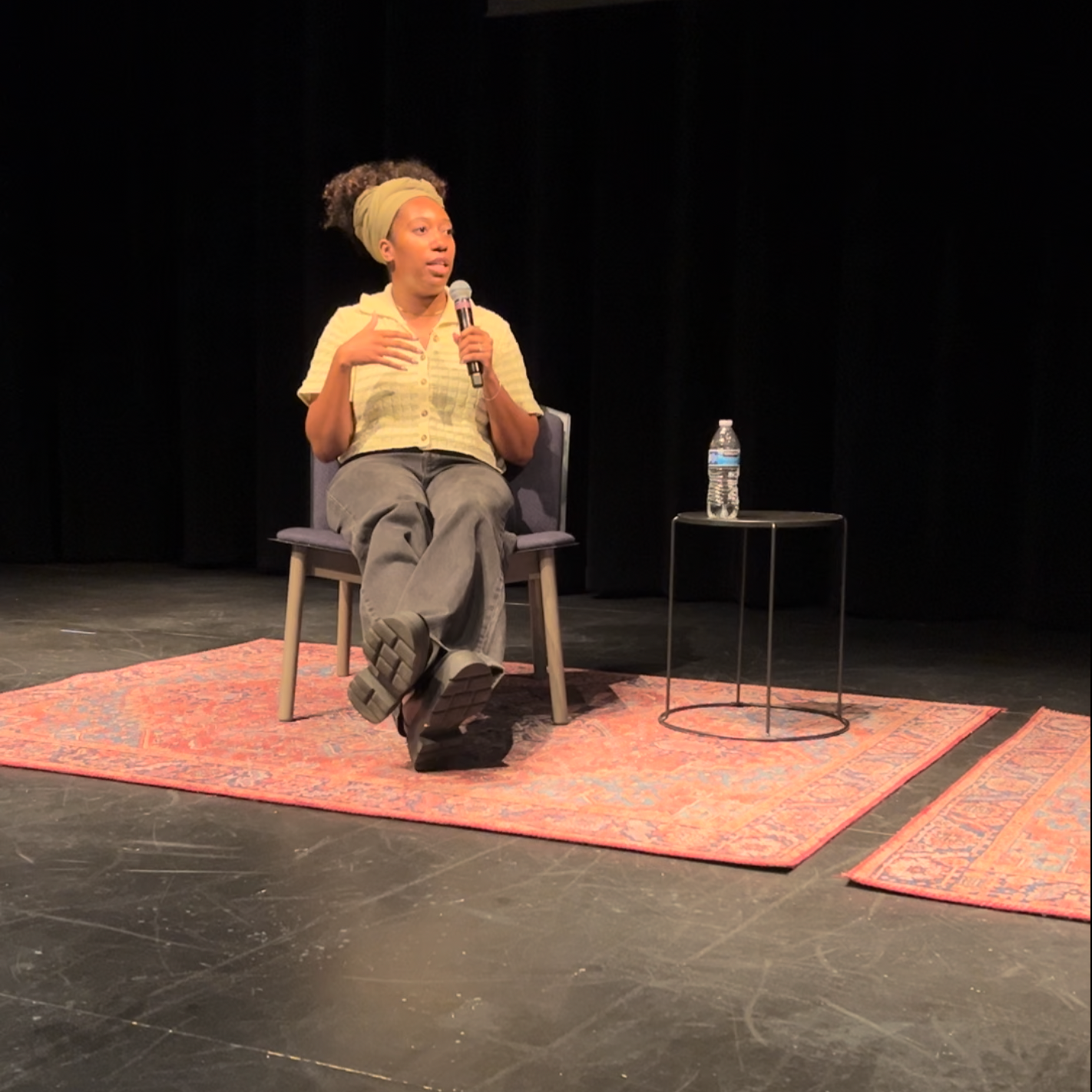 A woman sits on a chair on a stage, speaking into a microphone. There is a small table with a water bottle beside her, and she is sitting on a decorative rug.