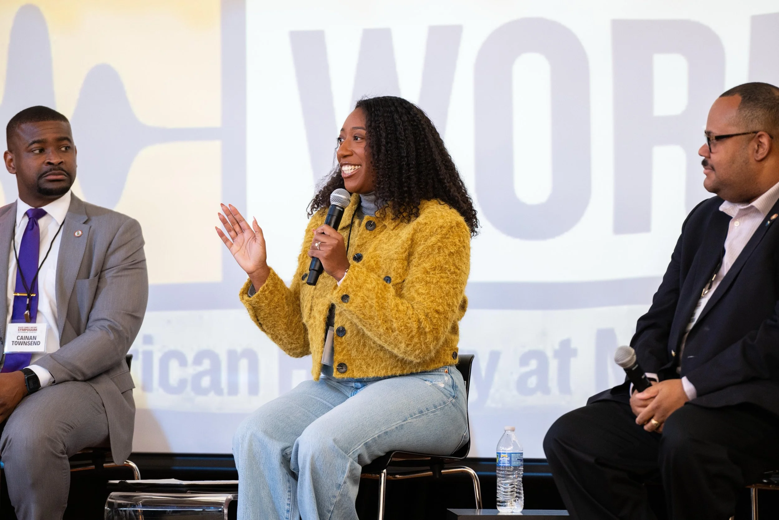 A woman in a yellow coat speaking into a microphone during a panel discussion, with two men seated beside her on stage.