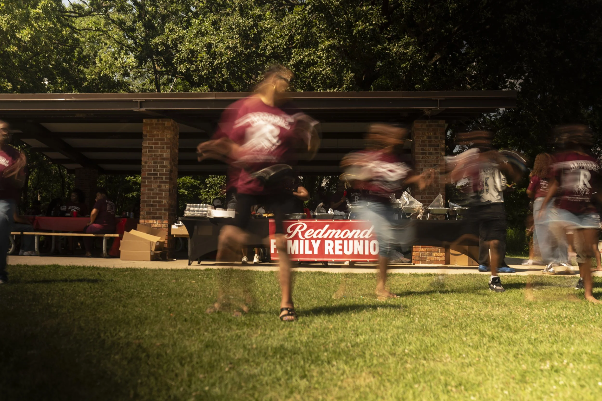 People walking past a table with a banner that reads 'Redmond Family Reunio' in a park with trees, some sitting at a picnic table, blurred motion of individuals in maroon and white clothing.