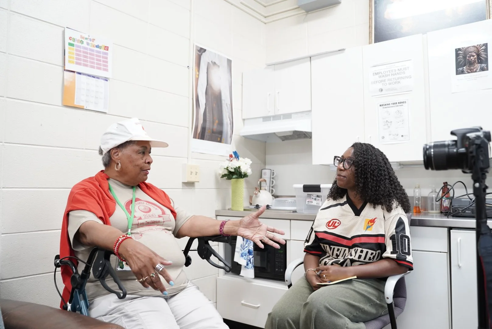 Two women having a conversation in a clinical setting, one wearing a wheelchair and the other sitting on a chair. The room has white walls, cabinets, and medical posters, with a camera on a tripod recording the discussion.