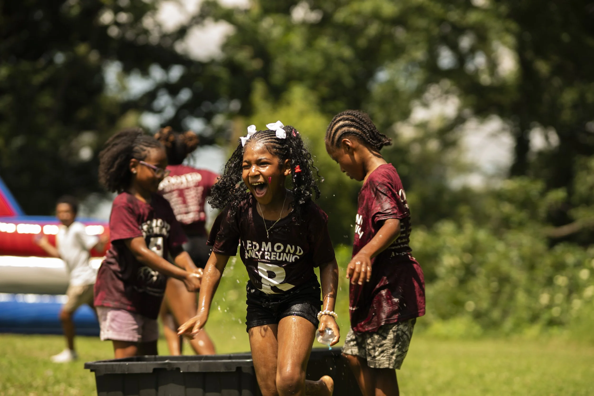 Children playing outdoors, with one girl in a black T-shirt smiling and others in maroon shirts, in a grassy area with trees in the background.