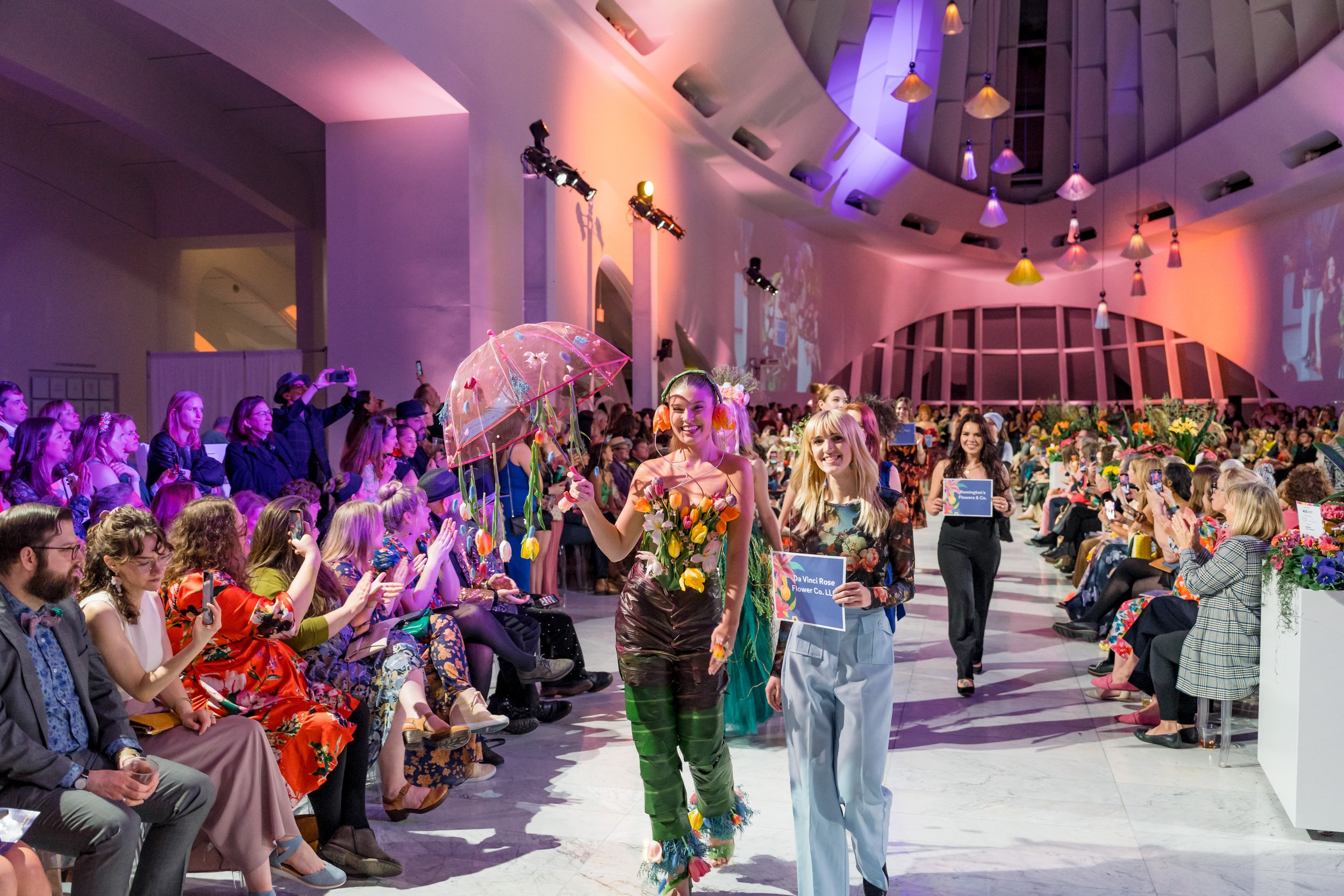 Fashion show featuring models walking down the runway with floral-themed outfits, including one holding a transparent umbrella with colorful decorations, in a well-lit, modern indoor venue with an audience seated on both sides taking pictures.