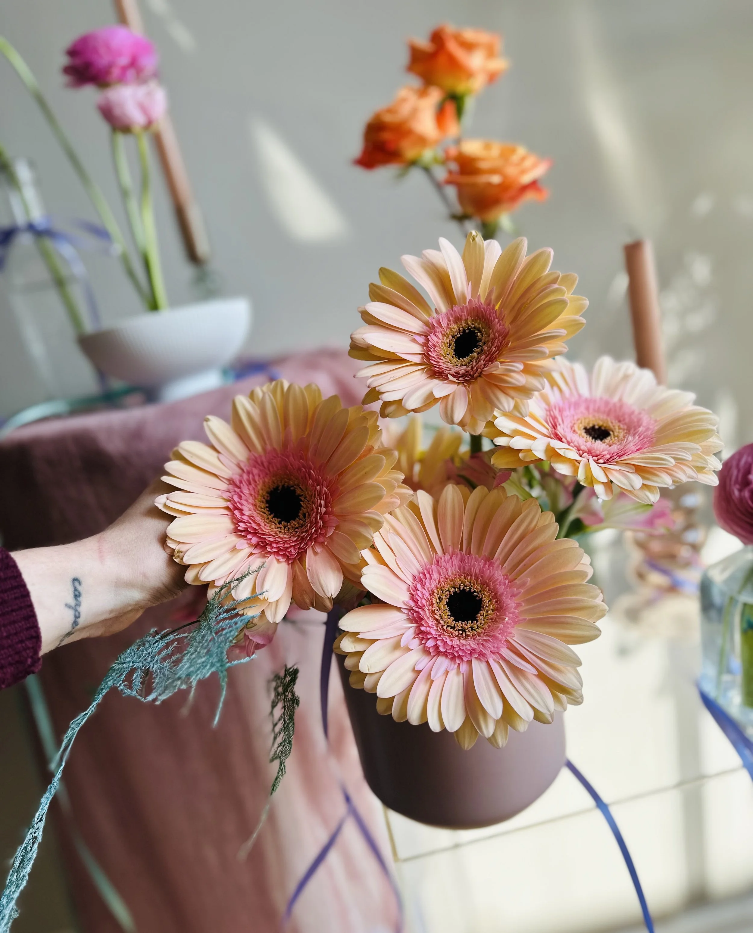 Close-up of a bouquet of pink and peach gerbera daisies being arranged indoors with other flowers and decorative items in the background.