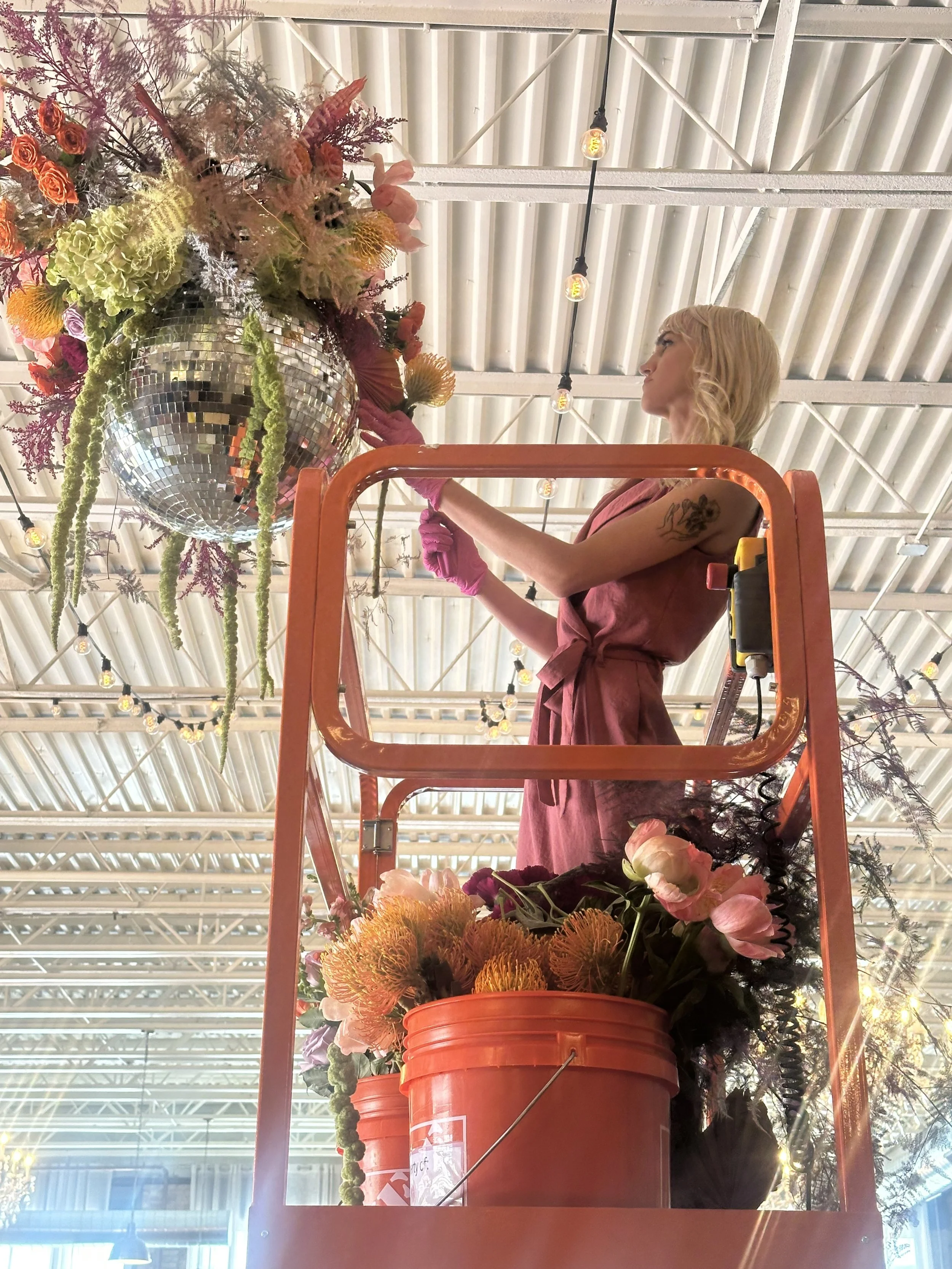 A woman with blonde hair on a lift, arranging a large floral display with a disco ball, various flowers, and greenery in an industrial-style space with string lights.