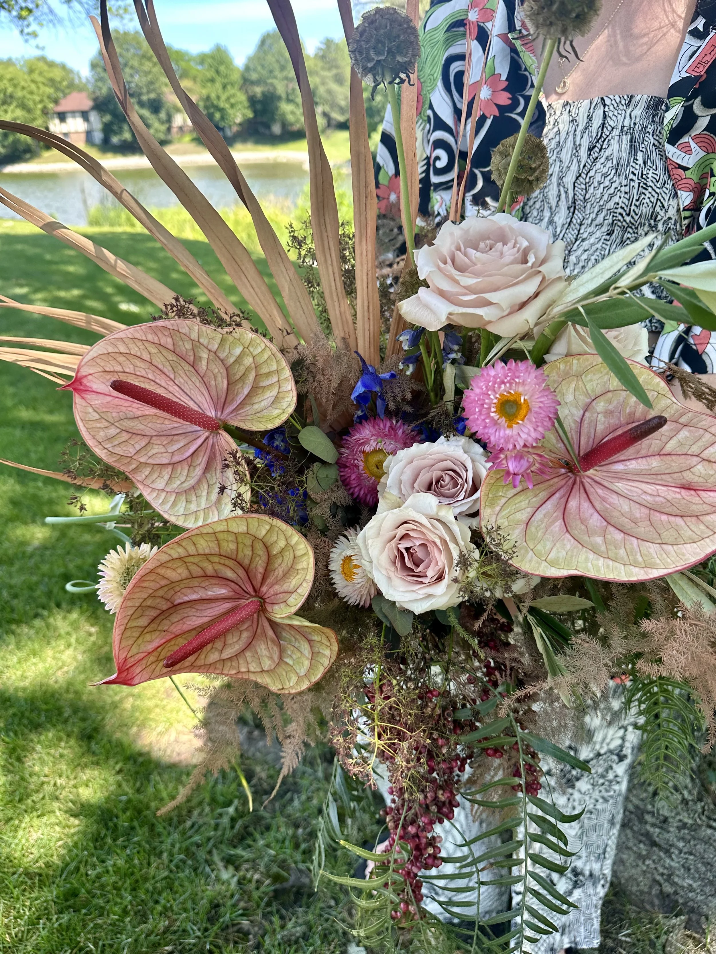 Colorful bouquet of flowers including pink roses, pink and white daisies, red anthuriums, and other greenery, held outdoors near a body of water with trees and houses in the background.