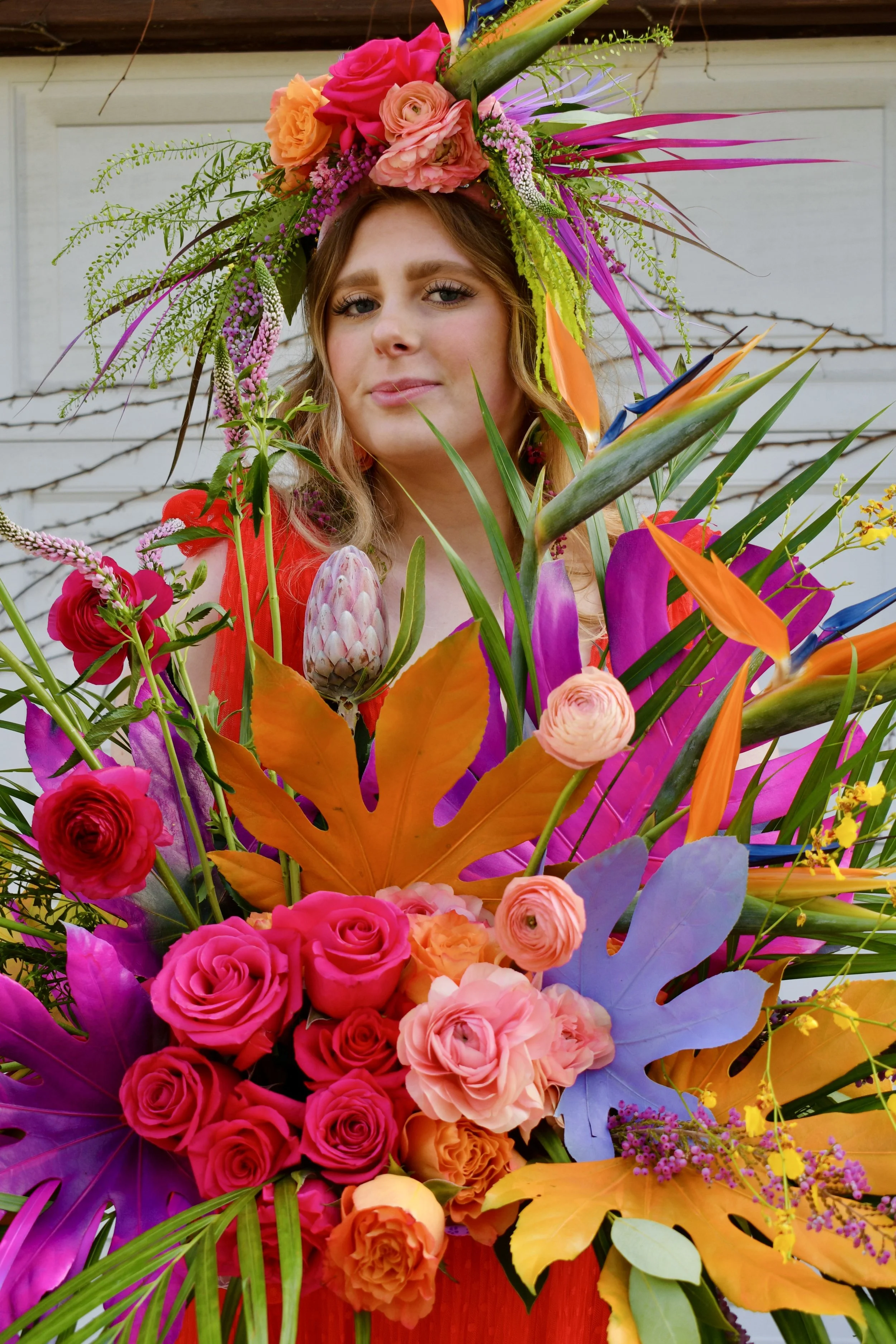 A woman with wavy blonde hair wearing a large flower crown, surrounded by a vibrant assortment of flowers and leaves.
