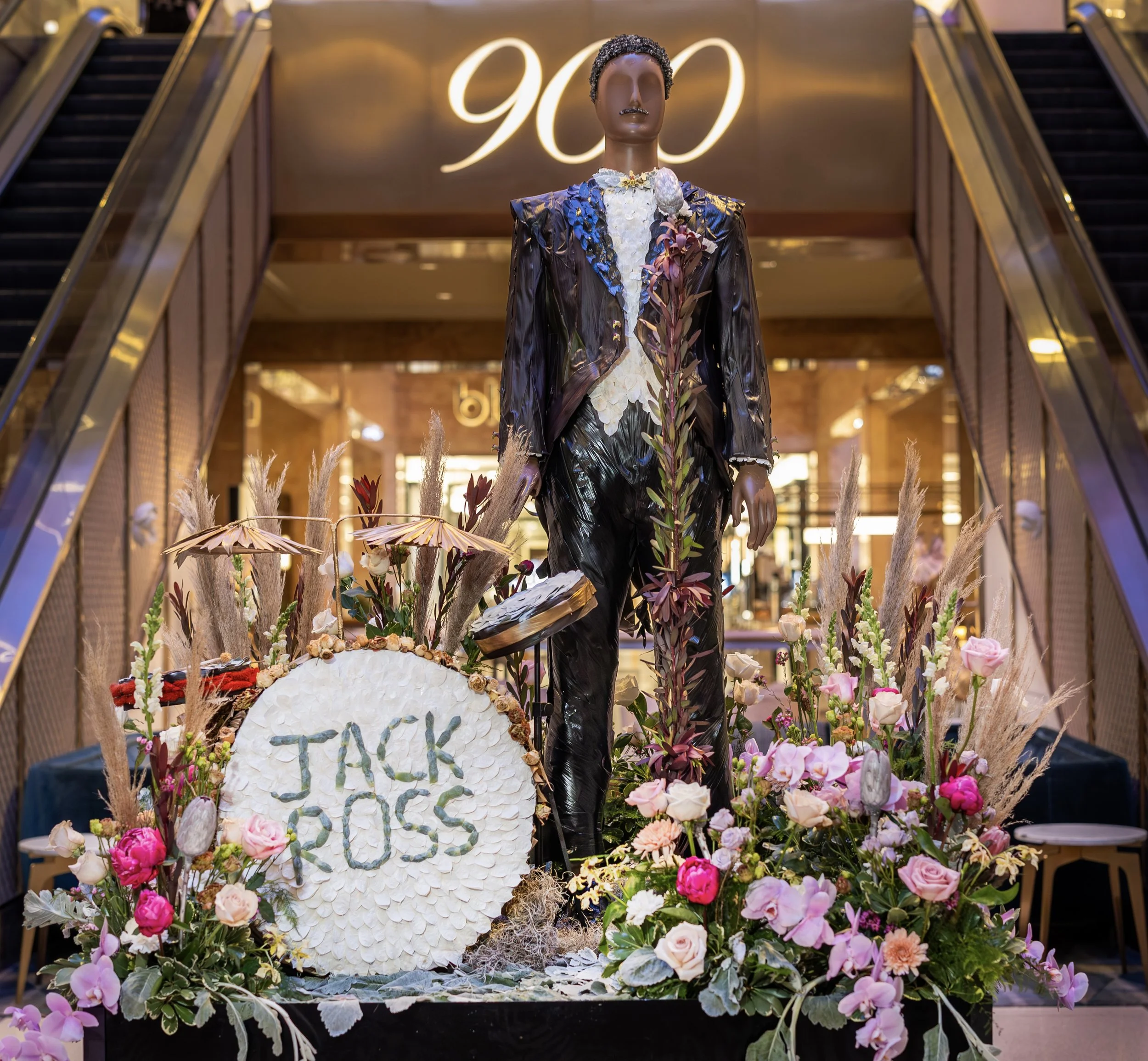 A decorative mannequin dressed in a floral and leather outfit stands on a floral display with a sign that reads 'JACK ROSS.' The display is surrounded by pink, white, and purple flowers, pampas grass, and decorative plants. The scene takes place at t