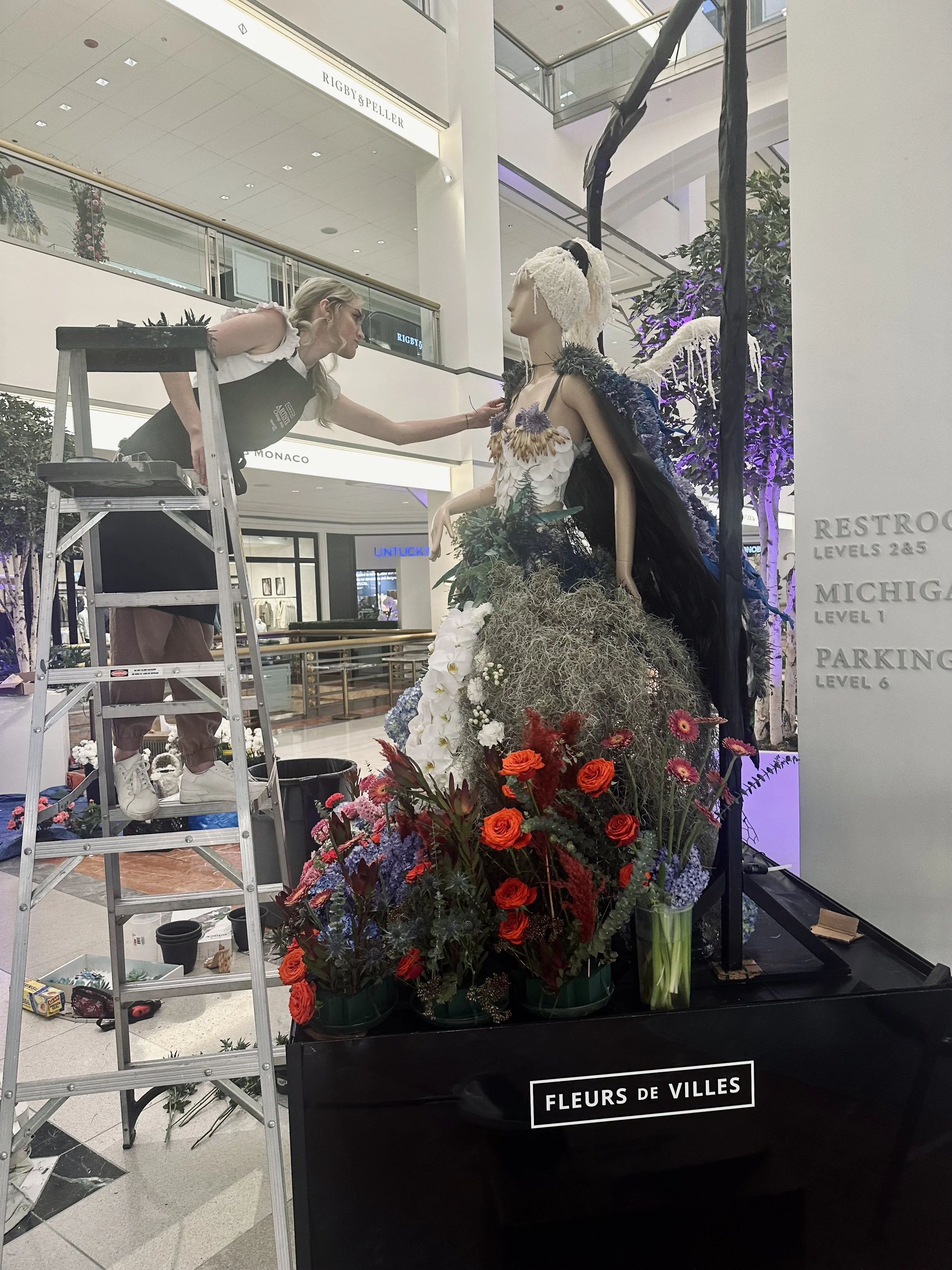 A florist arranging flowers on a mannequin display inside a shopping mall. The mannequin is dressed in an elaborate flower-themed outfit with various colorful flowers and greenery, and the scene is decorated with additional floral arrangements.