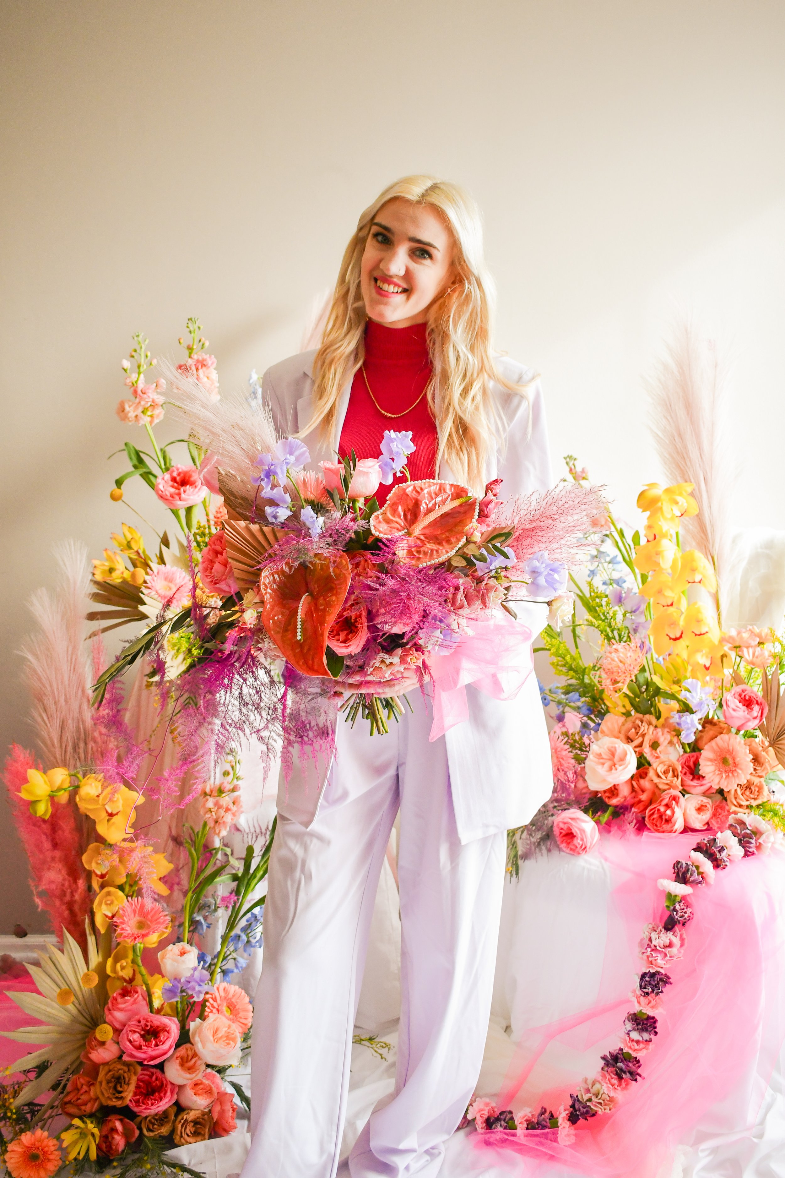 A woman with long blonde hair, wearing a red turtleneck and white blazer with white pants, holding a large colorful bouquet of flowers. She is standing in front of a display of vibrant flower arrangements, including pink, yellow, purple, peach, and o