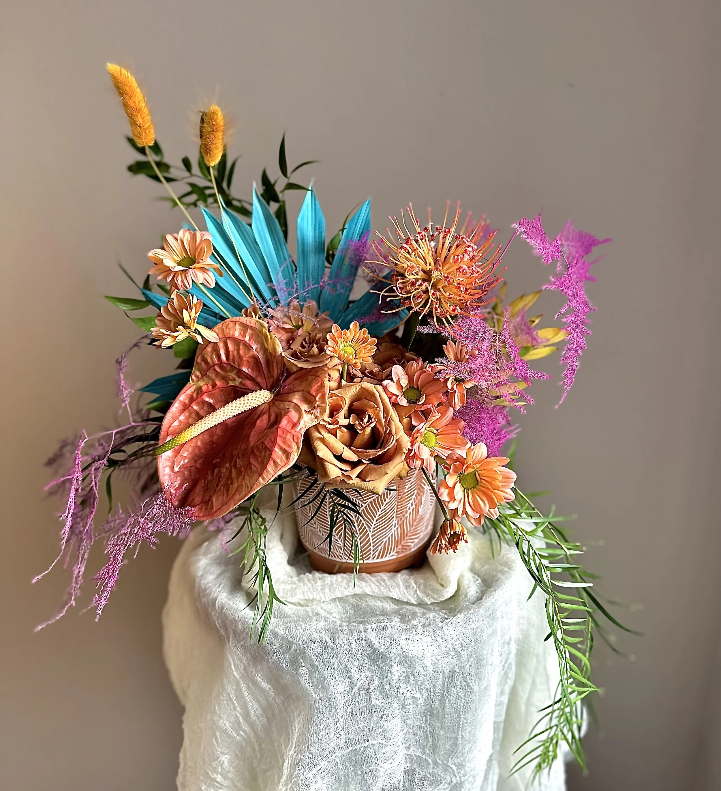 A colorful flower arrangement in a decorative pot on a white pedestal with a beige background.