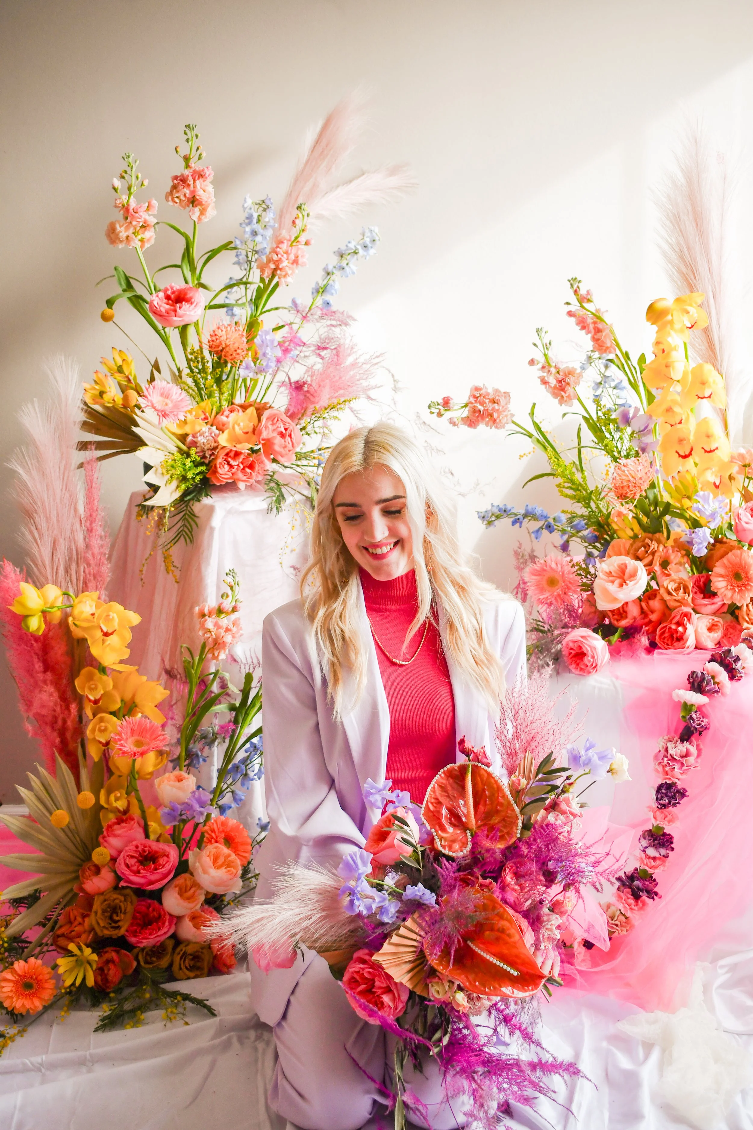 A woman with blonde hair, wearing a pink shirt and white blazer, smiling while surrounded by vibrant pink, purple, yellow, and orange flowers arranged in bouquets and displays.